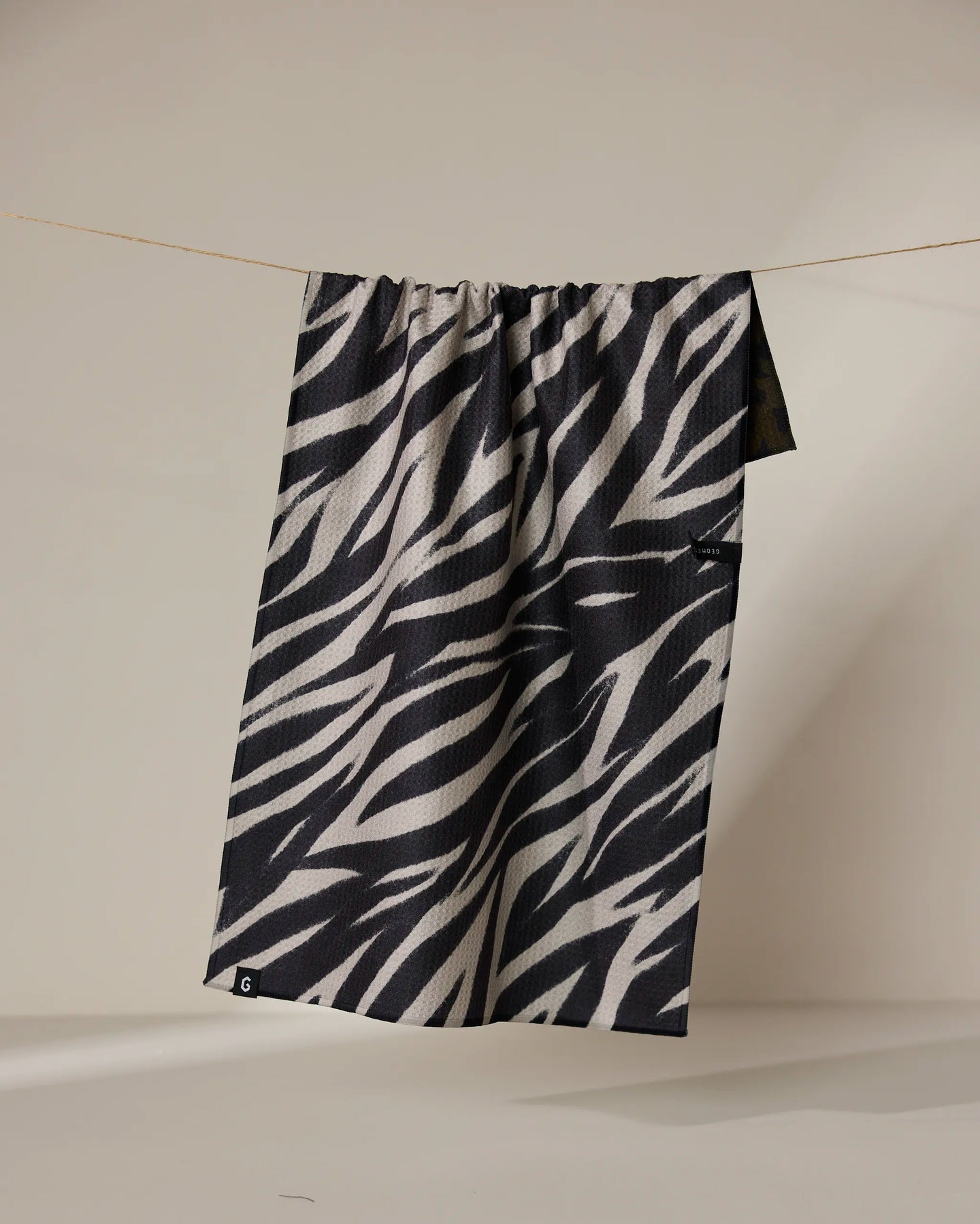 Geometry’s Safari tea towel, featuring a black and white zebra print and made with absorbent, quick-dry fabric, hangs on a clothesline against a plain beige background.