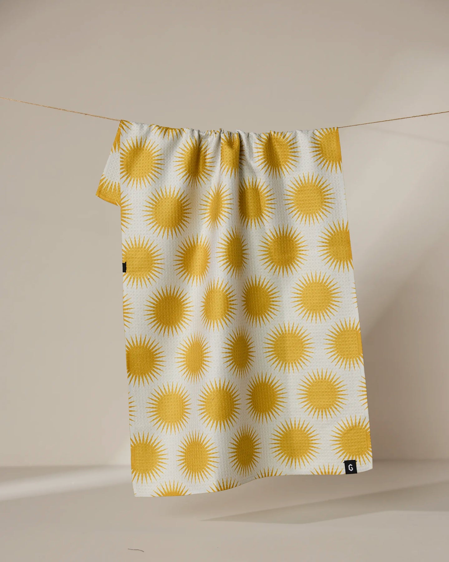 Geometry’s Energize recycled kitchen towel, featuring a repeating yellow sunburst pattern, hangs on a clothesline against a neutral background.