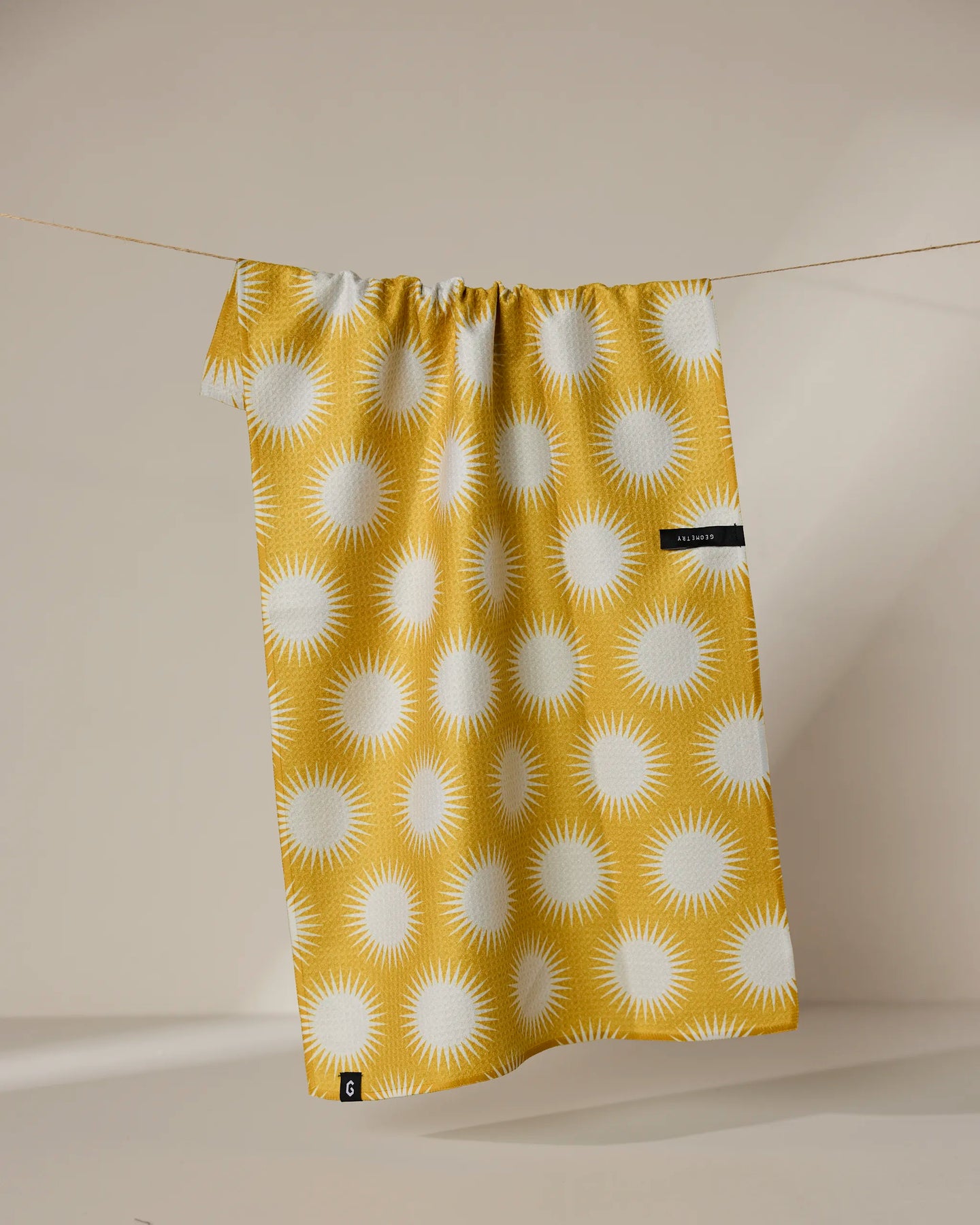 The Geometry Energize towel, featuring a white sunburst pattern on yellow and made from absorbent quick-dry material, hangs on a clothesline against a neutral beige background.