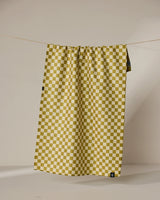 The Geometry Zebra Way tea towel in mustard yellow and white check pattern hangs from a thin string against a beige background, with a small black tag on its lower right corner.