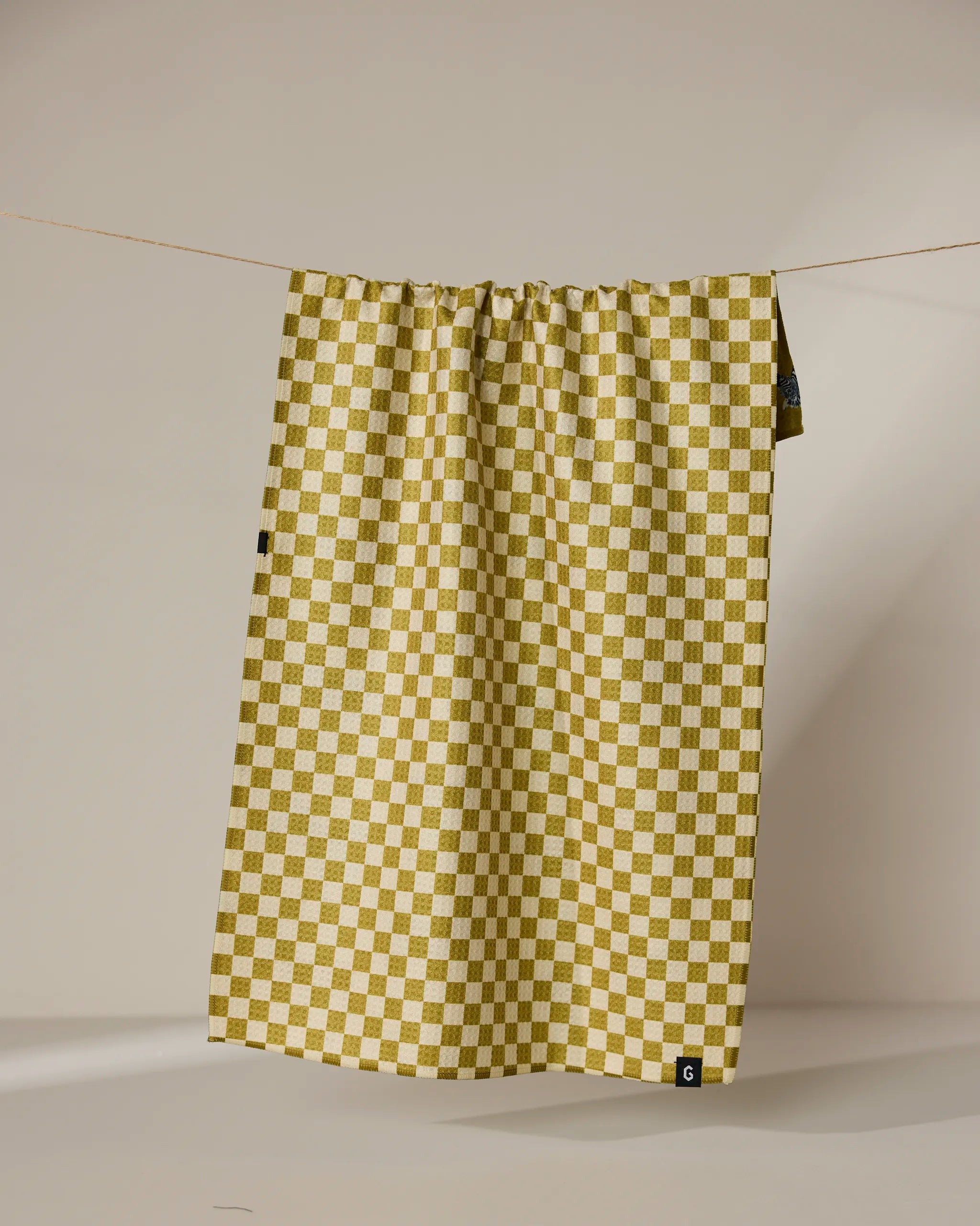 The Geometry Zebra Way tea towel in mustard yellow and white check pattern hangs from a thin string against a beige background, with a small black tag on its lower right corner.