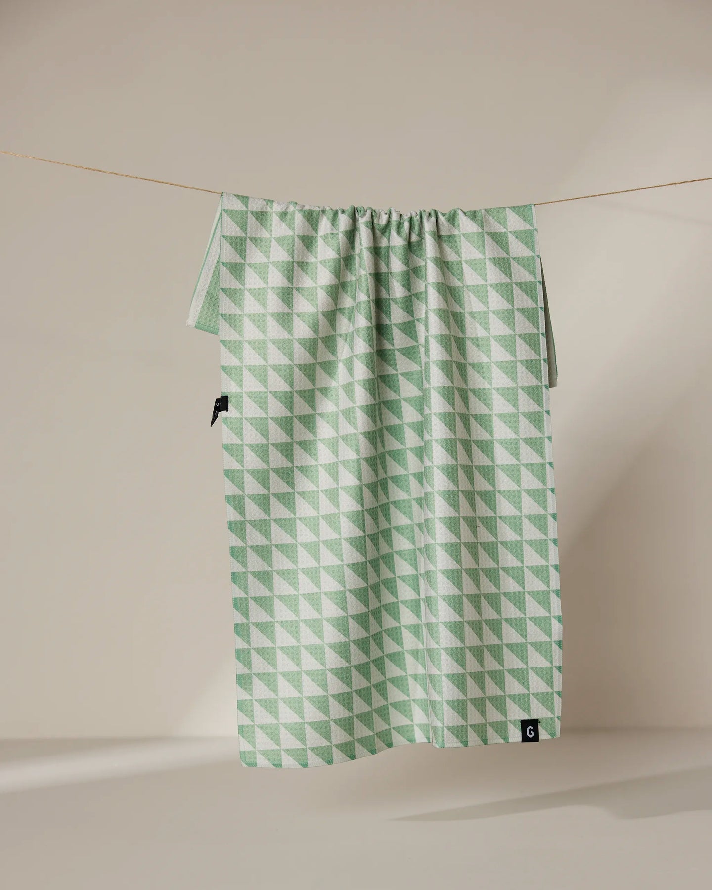 The Geometry Pistachio tea towel features a green and white double-sided geometric triangle pattern and hangs on a clothesline against a neutral background.