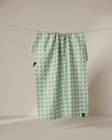 The Geometry Pistachio tea towel features a green and white double-sided geometric triangle pattern and hangs on a clothesline against a neutral background.