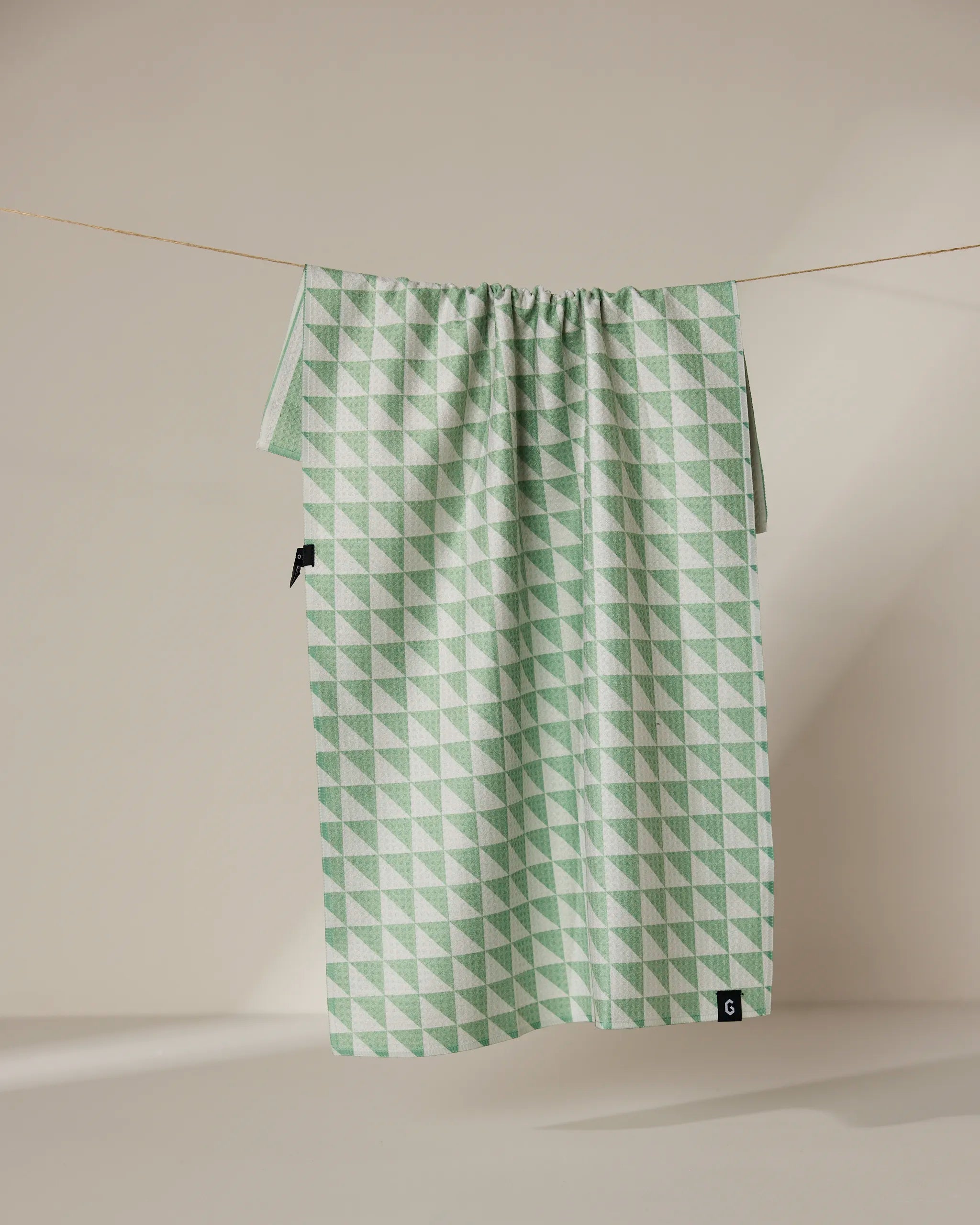 The Geometry Pistachio tea towel features a green and white double-sided geometric triangle pattern and hangs on a clothesline against a neutral background.