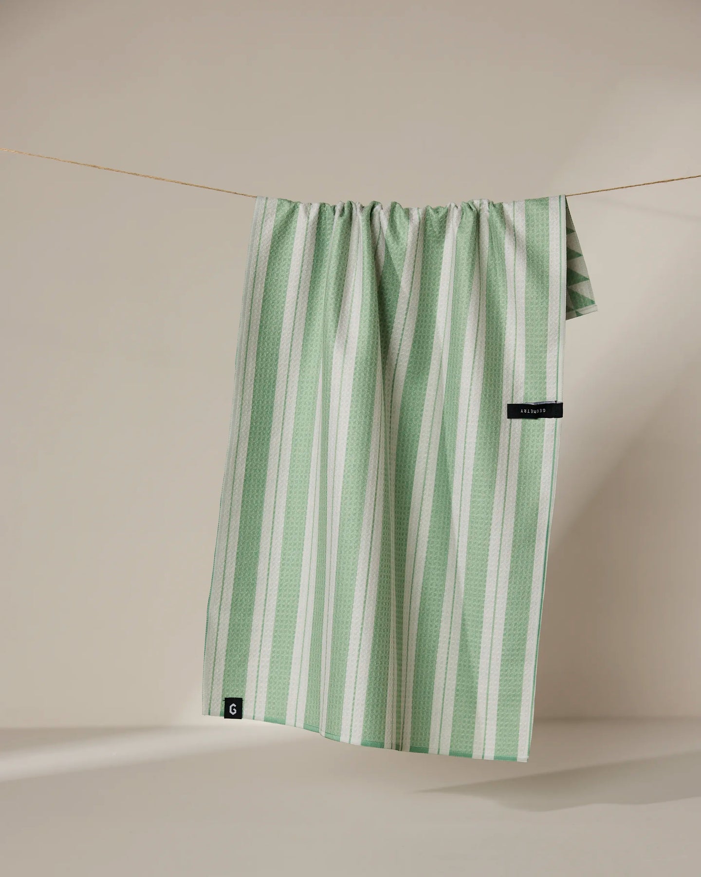 The Geometry Pistachio tea towel, featuring green and white stripes, hangs neatly over a thin, neutral-colored clothesline against a beige background.