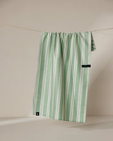 The Geometry Pistachio tea towel, featuring green and white stripes, hangs neatly over a thin, neutral-colored clothesline against a beige background.