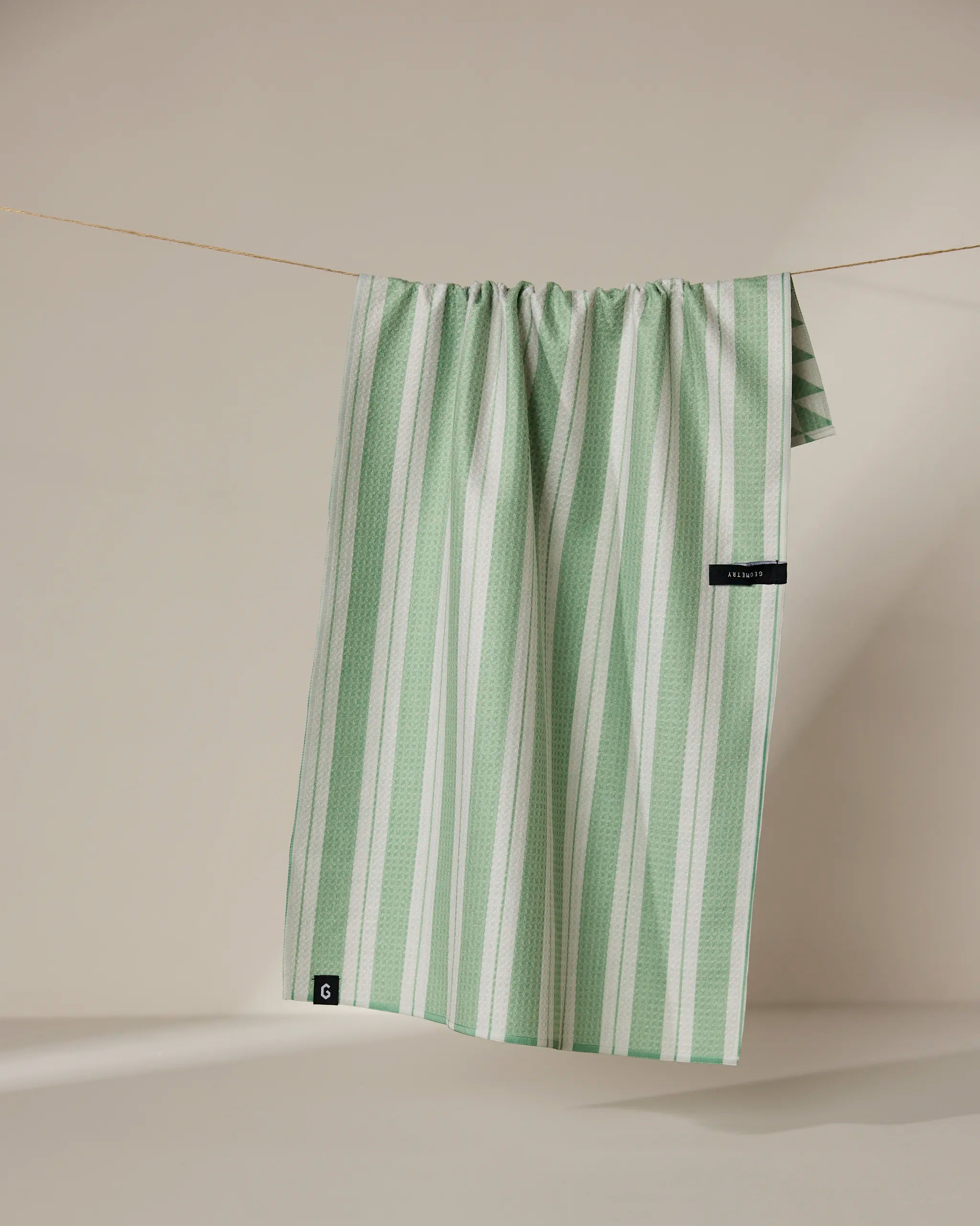 The Geometry Pistachio tea towel, featuring green and white stripes, hangs neatly over a thin, neutral-colored clothesline against a beige background.
