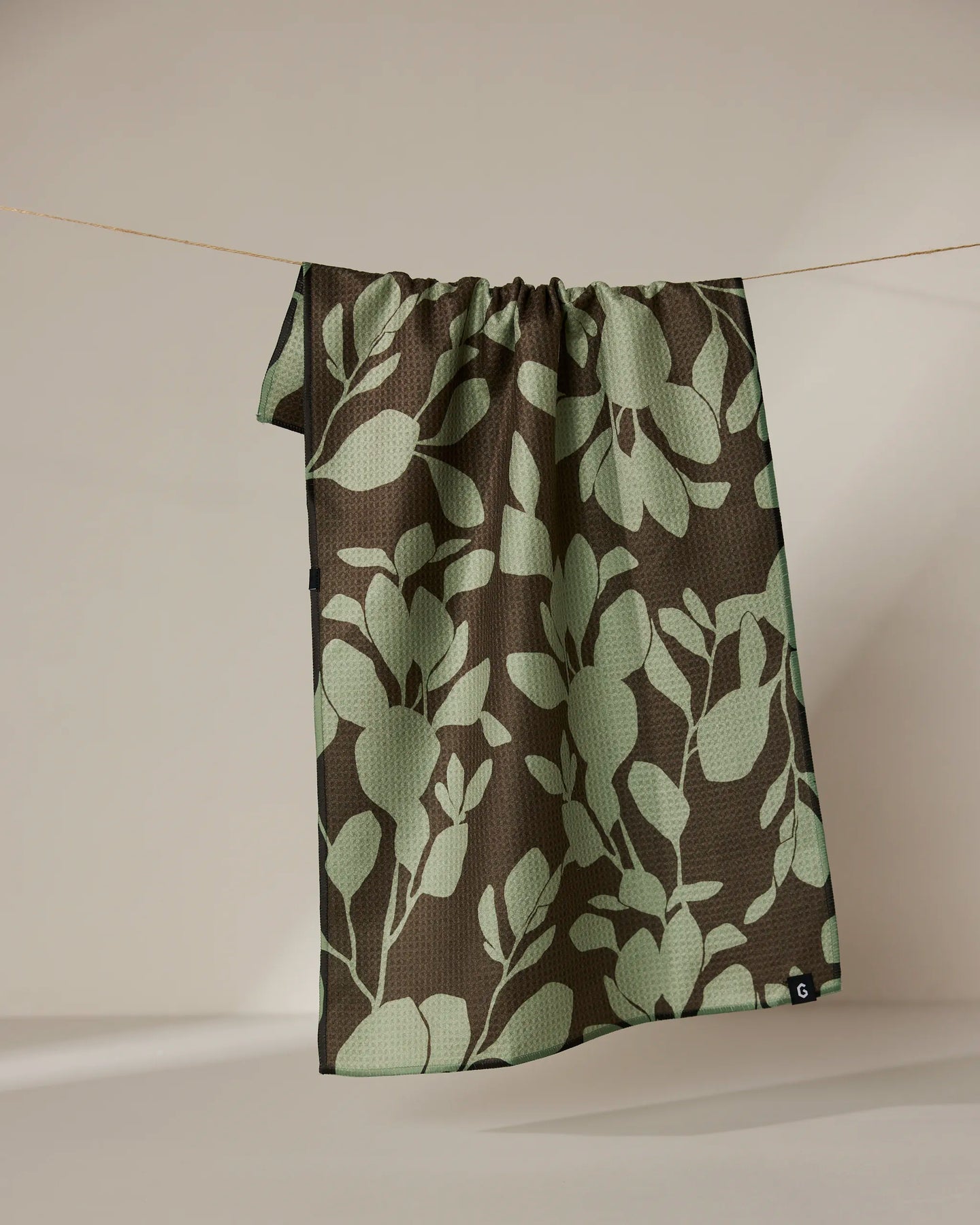 The Geometry Eucalyptus absorbent quick-dry towel, featuring a green leafy design on a dark brown background, hangs on a clothesline against a plain beige backdrop.