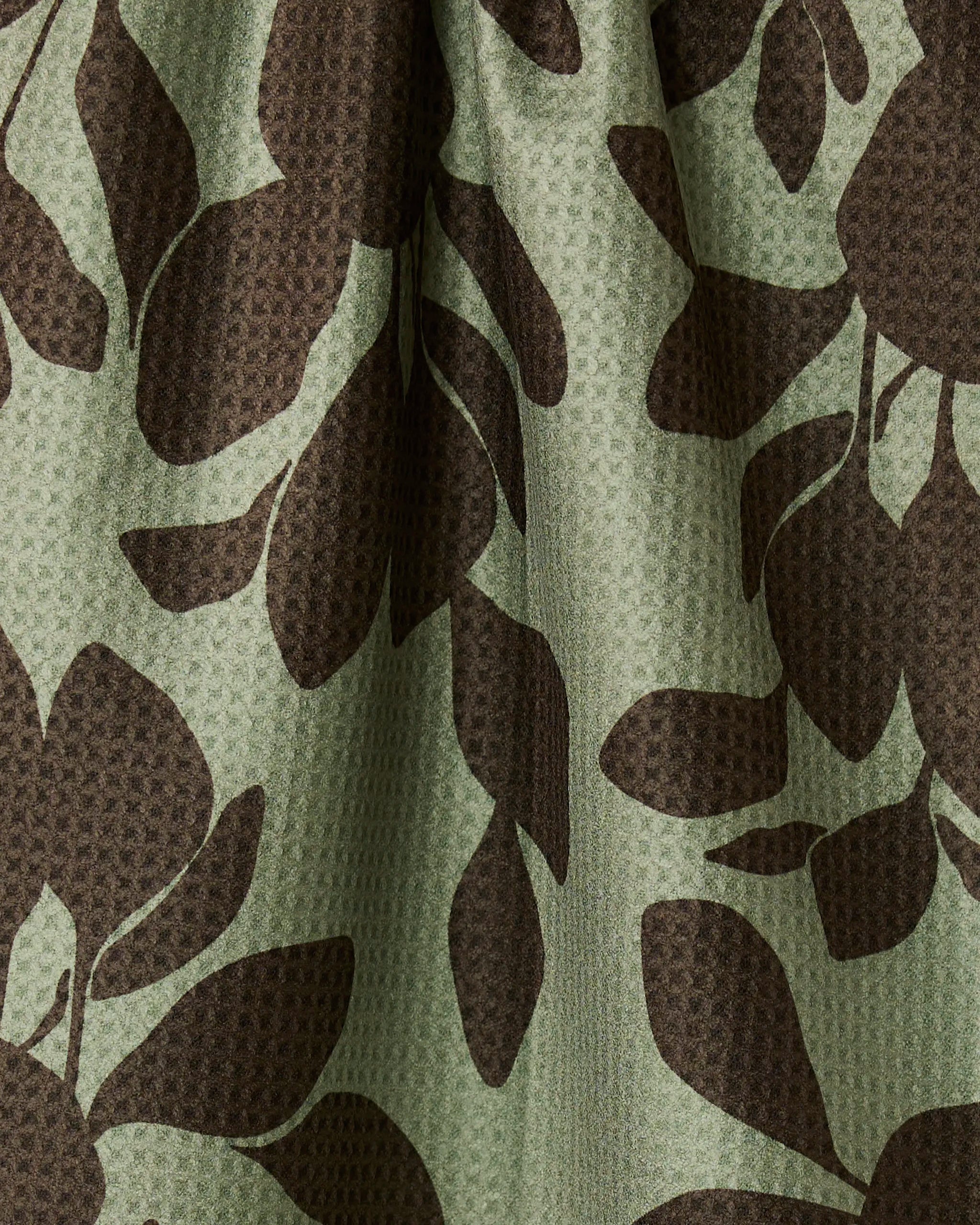 Close-up of Geometry’s Eucalyptus recycled kitchen towel featuring a green and dark brown leaf pattern. The textured, waffle-like fabric has soft folds that create subtle shadows and highlights.
