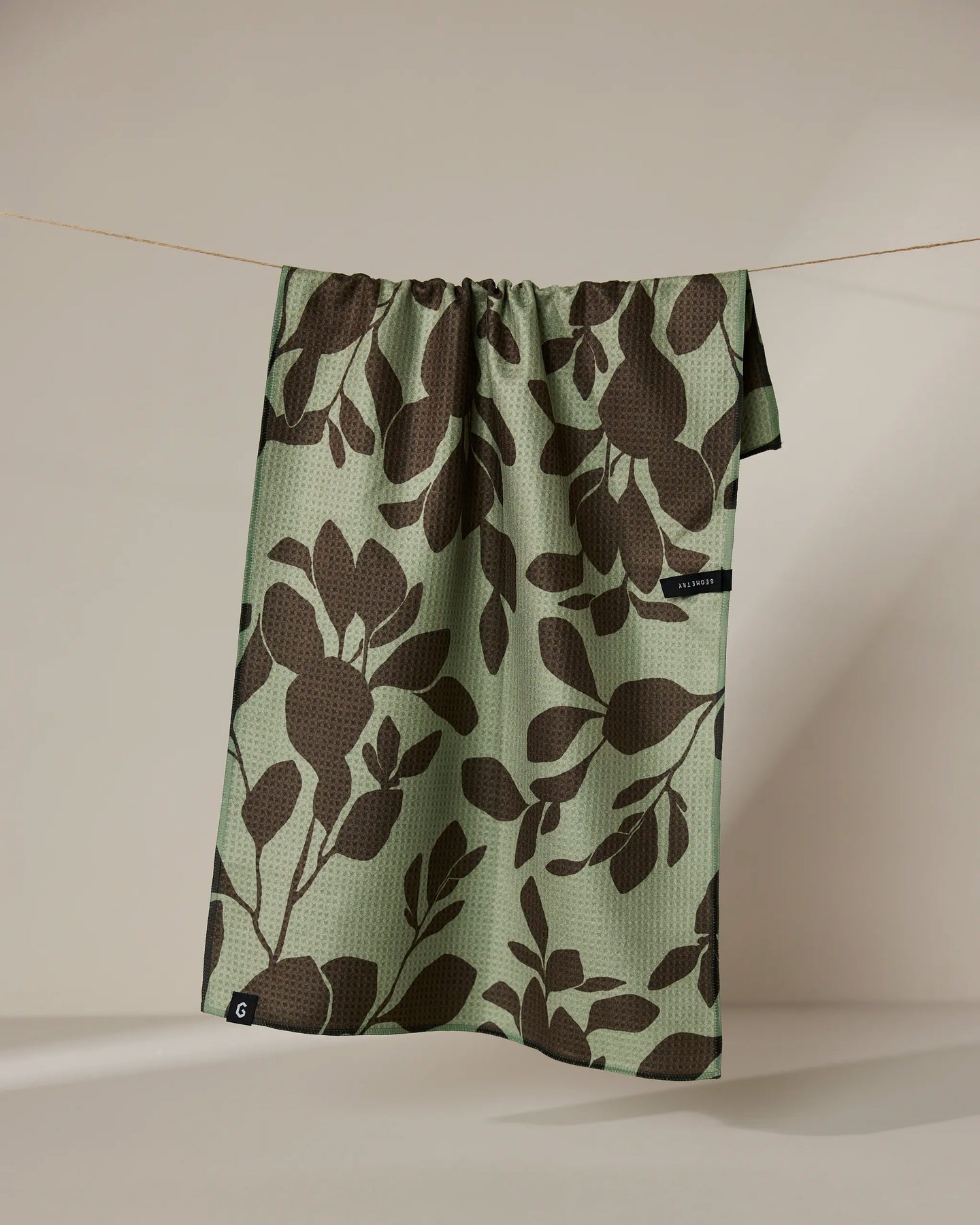 The Geometry Eucalyptus towel, featuring large dark brown leafy patterns on green and a small black tag at the edge, hangs on a string against a neutral background.