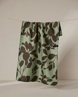 The Geometry Eucalyptus towel, featuring large dark brown leafy patterns on green and a small black tag at the edge, hangs on a string against a neutral background.