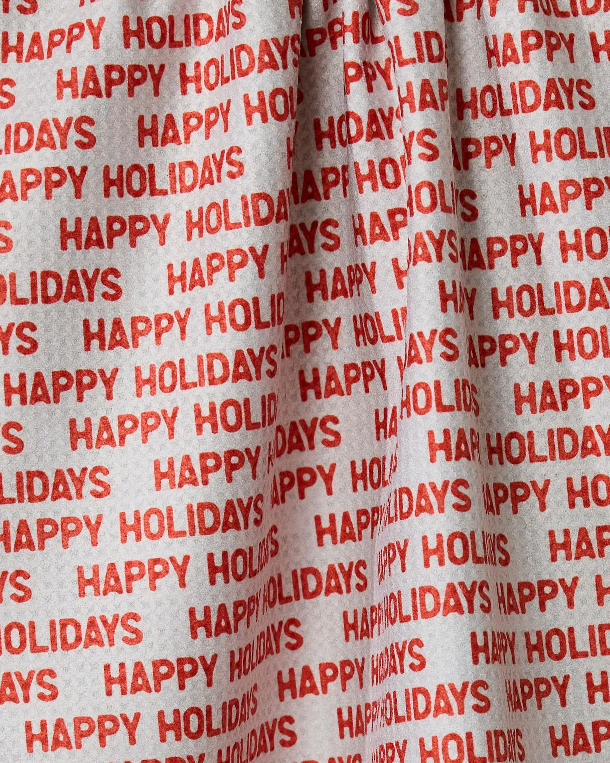 The Geometry Holiday 2024 towel is made of white fabric with repeated red HAPPY HOLIDAYS text in all caps, arranged in horizontal rows. Absorbent and quick-drying, its festive look is enhanced by the gathered or folded sections.