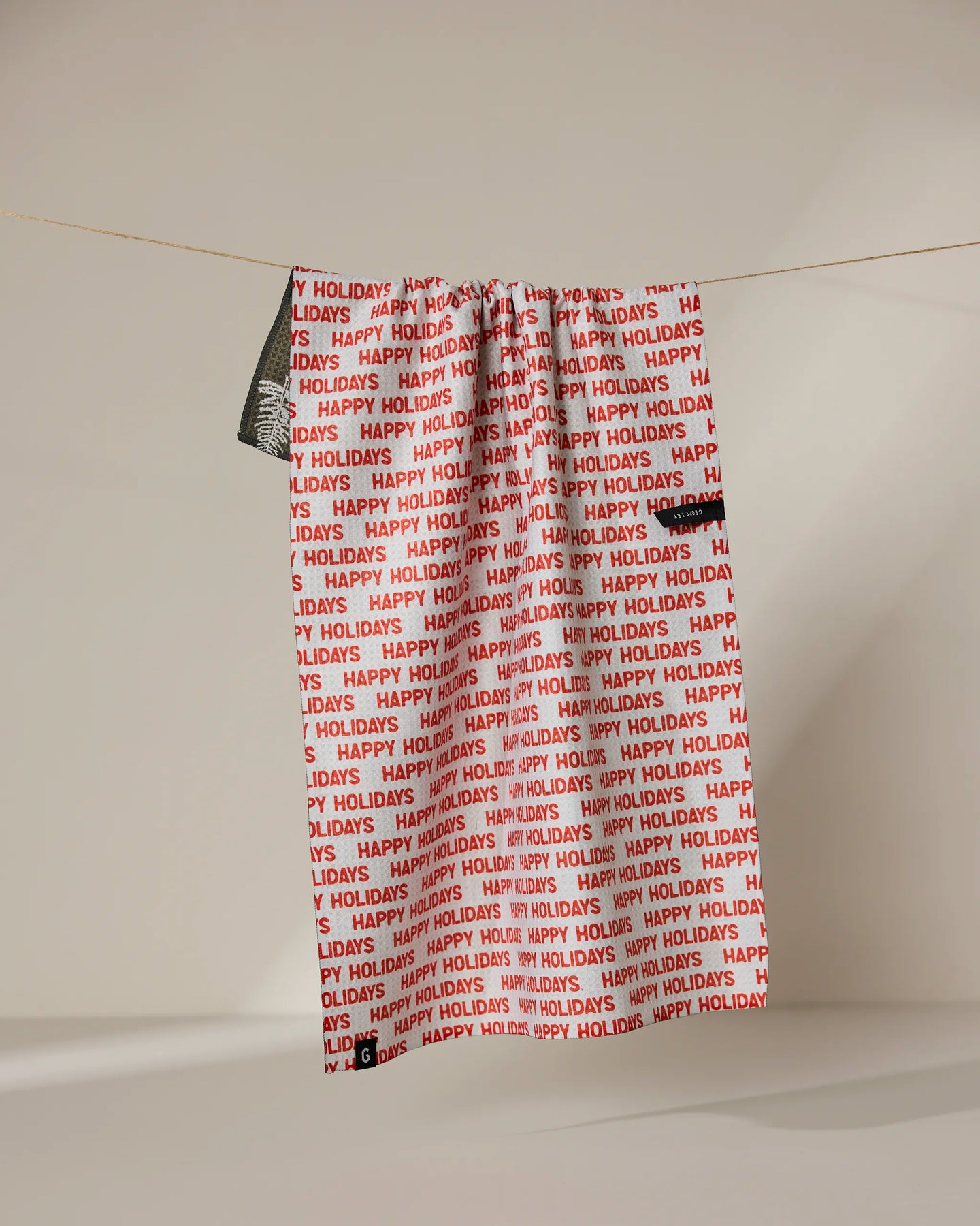 A Geometry Holiday 2024 gift bag featuring red and white HAPPY HOLIDAYS print hangs beside an absorbent, quick-drying towel—ideal for festive kitchen cleanup—against a neutral background.