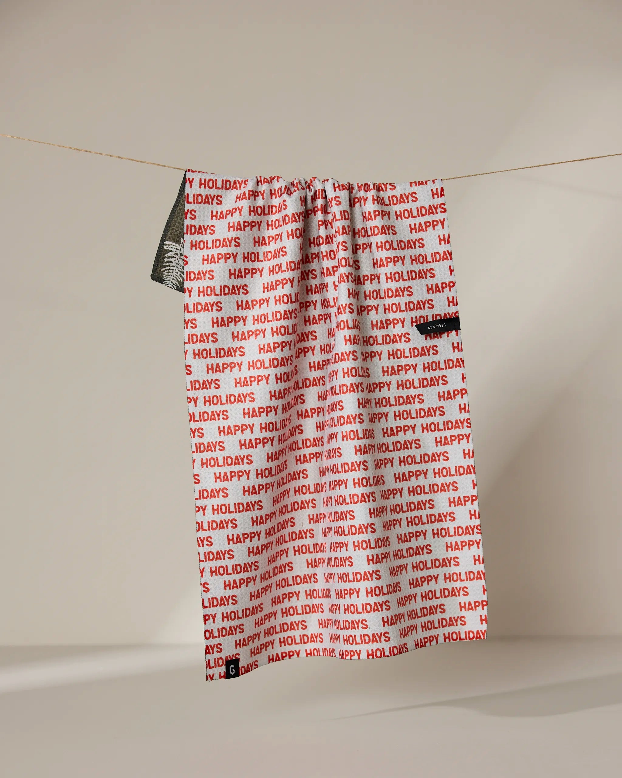 A Geometry Holiday 2024 gift bag featuring red and white HAPPY HOLIDAYS print hangs beside an absorbent, quick-drying towel—ideal for festive kitchen cleanup—against a neutral background.