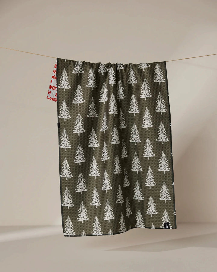 A green recycled "Holiday" kitchen towel by Geometry, featuring white pine trees and a red and white tag, hangs on a clothesline against a neutral background. This absorbent, quick-dry towel is eco-friendly and stylish.