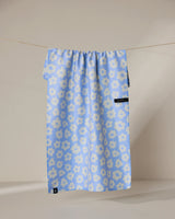 The Peri recycled kitchen towel by Geometry, featuring a blue design with white floral patterns, hangs on a clothesline against a neutral background. Small black tags appear near the bottom and right side of the towel.