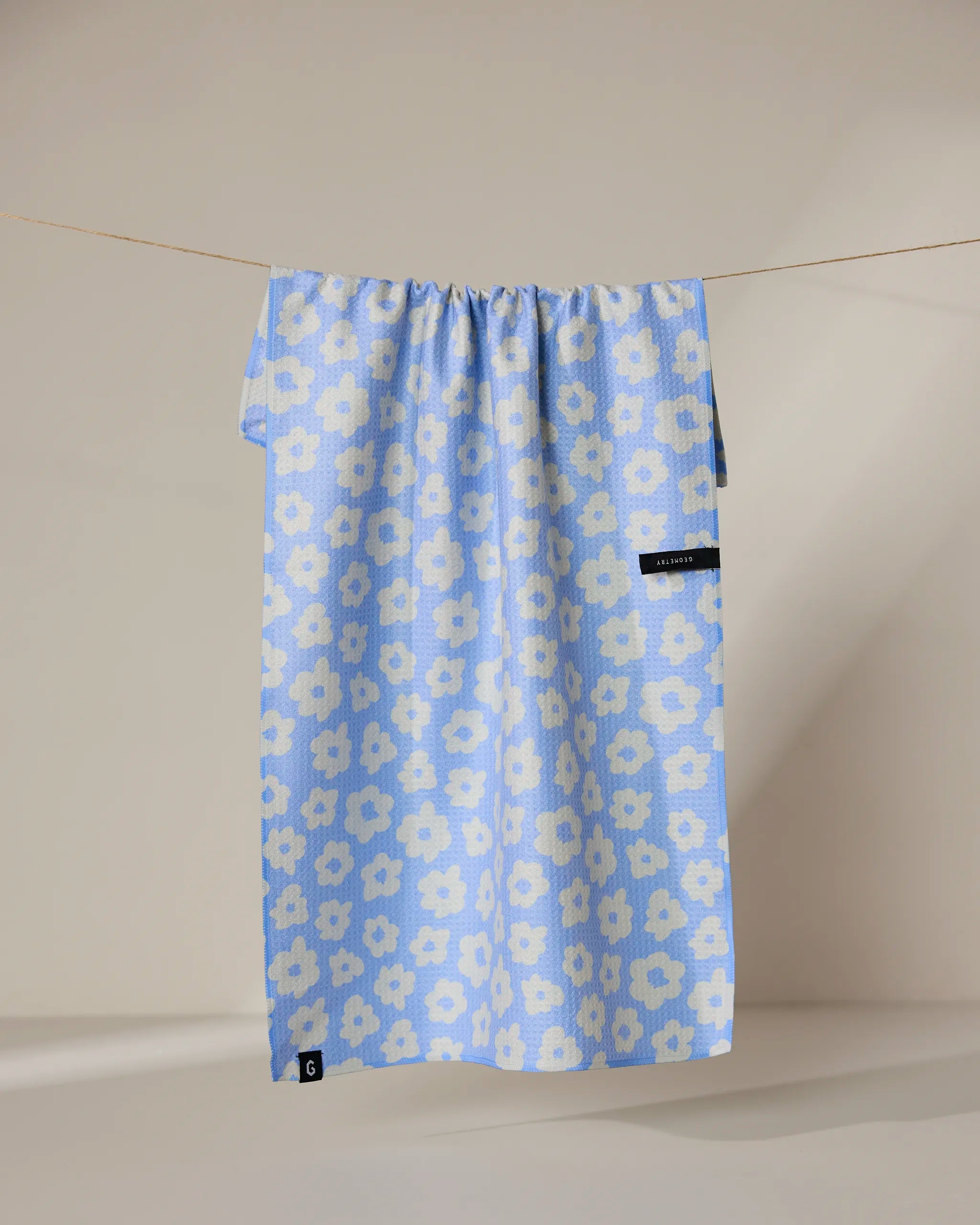 The Peri recycled kitchen towel by Geometry, featuring a blue design with white floral patterns, hangs on a clothesline against a neutral background. Small black tags appear near the bottom and right side of the towel.
