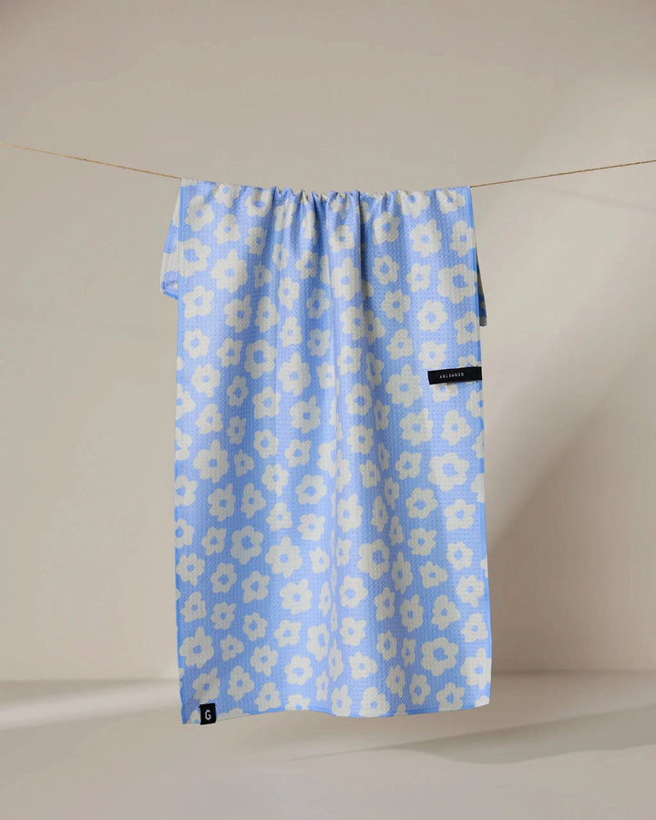 The Peri recycled kitchen towel by Geometry, featuring a blue design with white floral patterns, hangs on a clothesline against a neutral background. Small black tags appear near the bottom and right side of the towel.