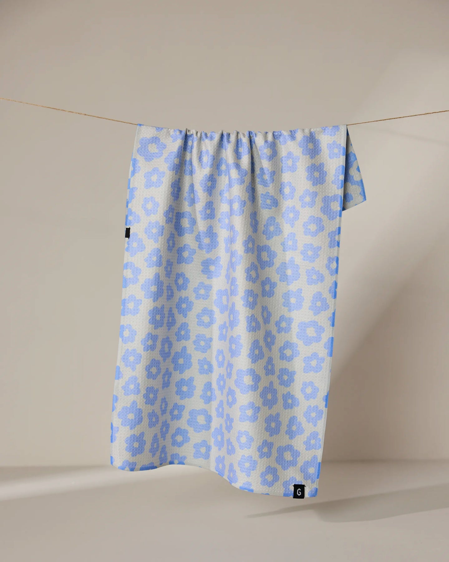 The Geometry Peri tea towel, featuring a light blue and white floral pattern, hangs neatly from a thin clothesline against a neutral beige background. Its quick-drying fabric combines style and function.