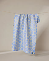 The Geometry Peri tea towel, featuring a light blue and white floral pattern, hangs neatly from a thin clothesline against a neutral beige background. Its quick-drying fabric combines style and function.