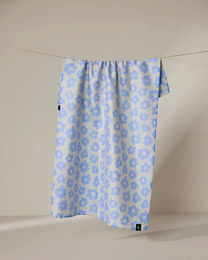 The Geometry Peri tea towel, featuring a light blue and white floral pattern, hangs neatly from a thin clothesline against a neutral beige background. Its quick-drying fabric combines style and function.