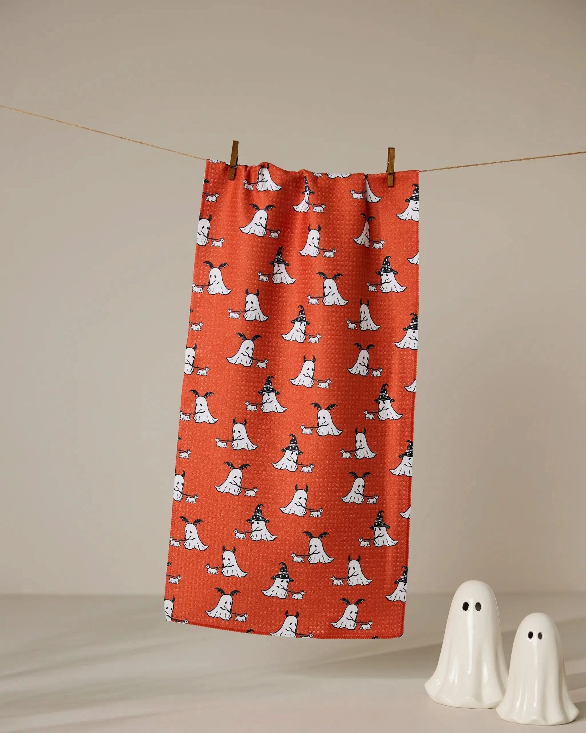 The Geometry Spooky Ghost Walk Orange ultra-absorbent tea towel features white cartoon ghosts in witch hats and hangs on a clothesline. Two small white ceramic ghost figurines are displayed below.