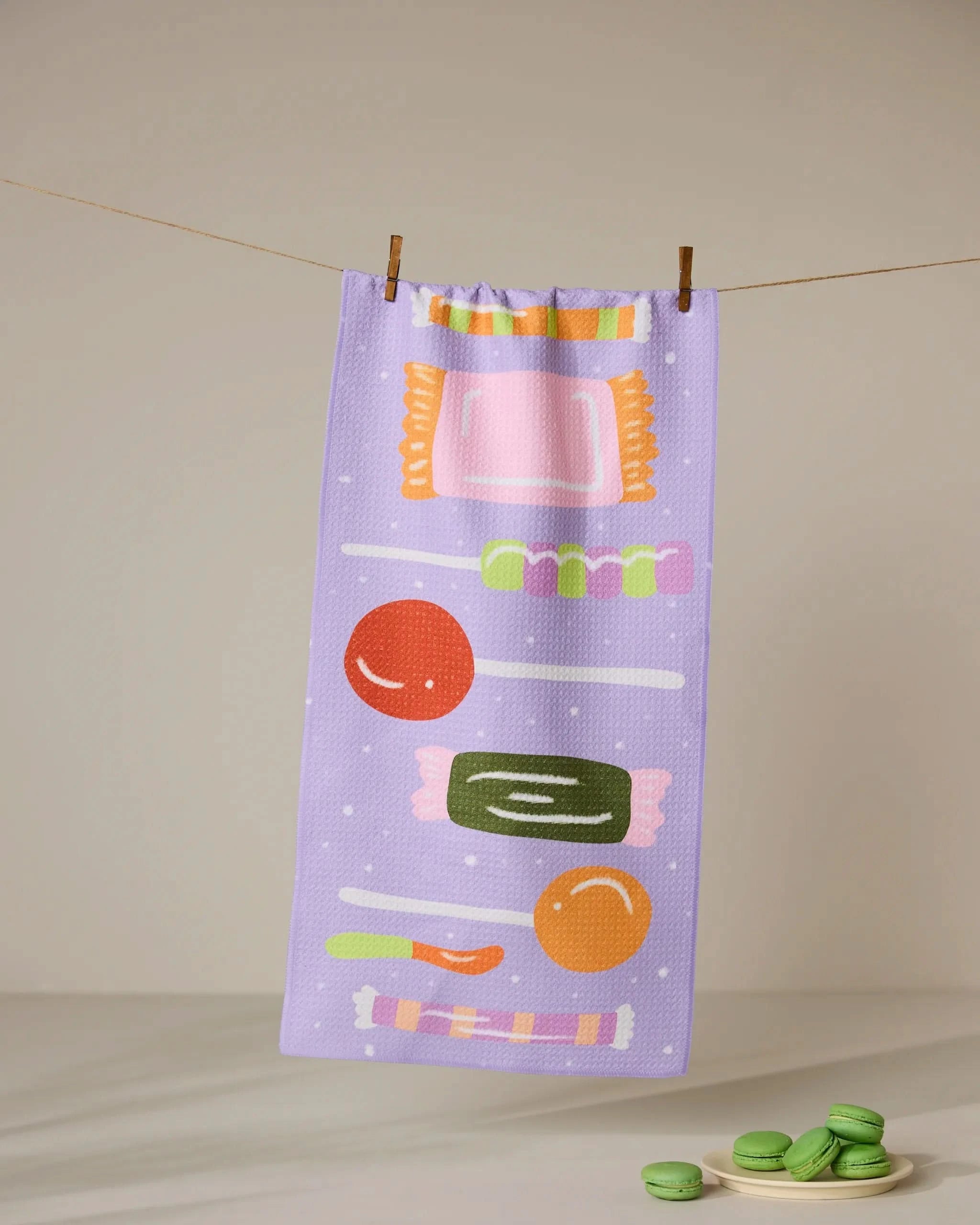 The Geometry Stacked Sweet Treasures towel in lavender, adorned with colorful illustrations of sweets—wrapped candies, a cookie, and a biscuit—hangs from a clothesline. Three green macarons are arranged below.