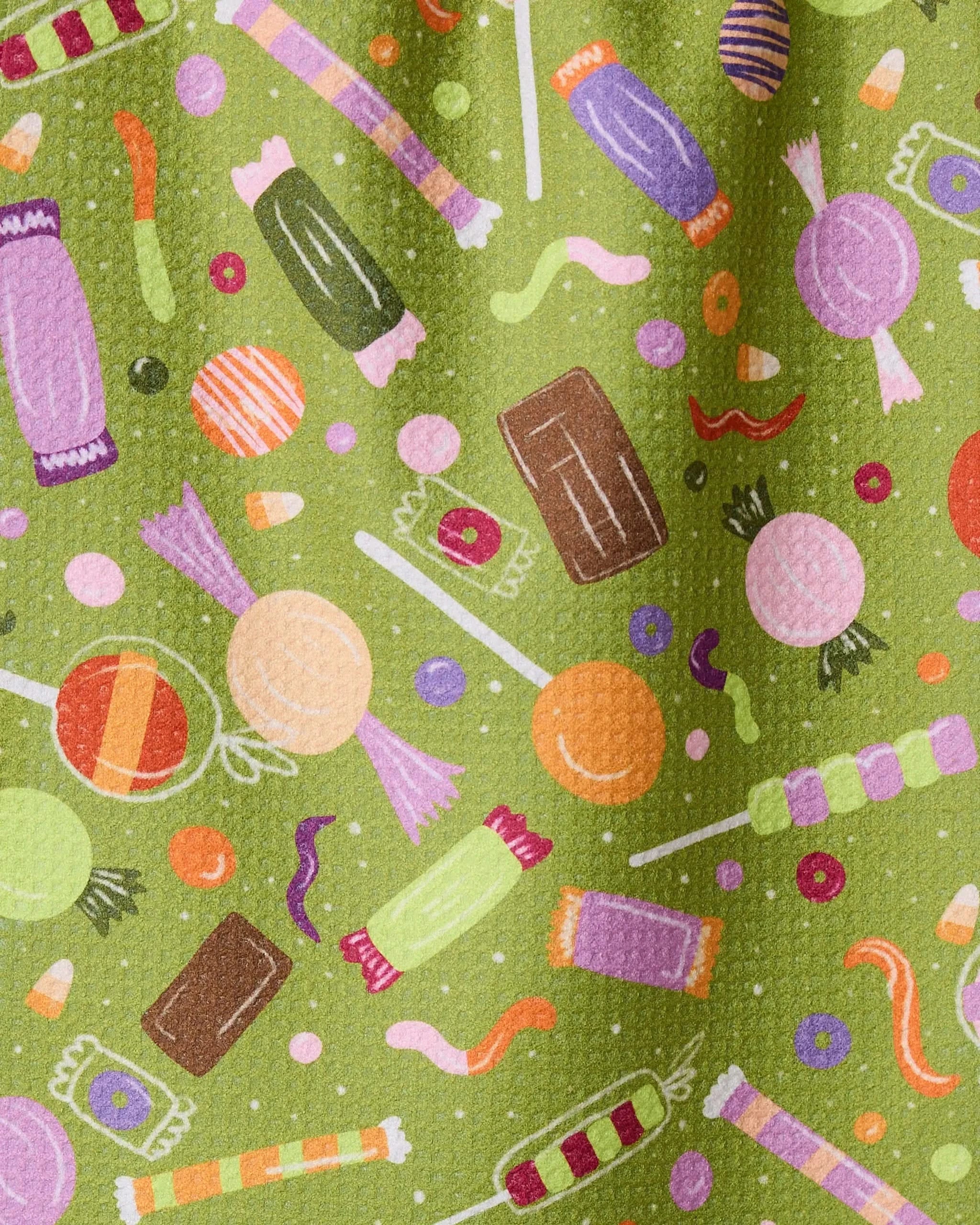 Sweet Treasures by Geometry features a colorful candy illustration—lollipops, wrapped chocolates, and gummy worms—scattered on a green Geoweave bar towel made from recycled materials.