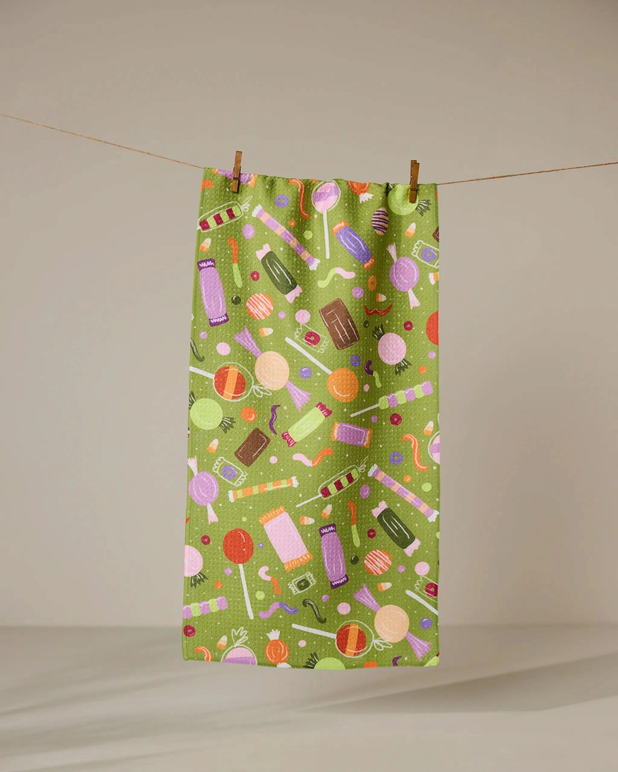 The Geometry Sweet Treasures bar towel features a colorful candy pattern with lollipops and chocolates on green, hanging against a beige background. Made from ultra-absorbent fabric, its perfect for kitchen use.