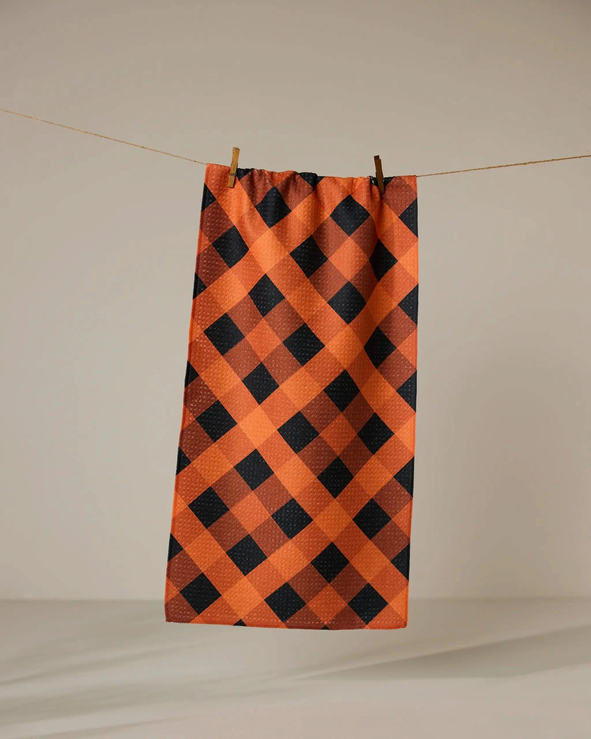 The Geometry Pumpkin Plaid quick-drying rectangular towel, featuring an orange and black plaid pattern, hangs from a clothesline with two wooden clothespins against a neutral background.