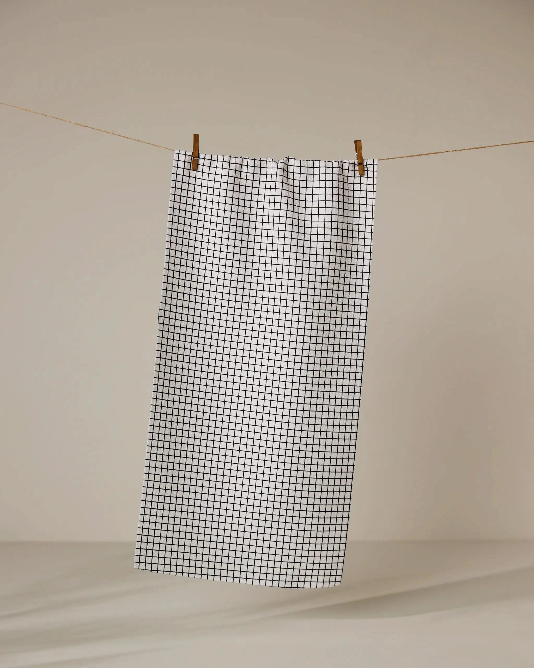 The Ian towel by Geometry, featuring a black grid pattern on ultra-absorbent white fabric, hangs on a clothesline with two wooden clothespins against a plain beige background.