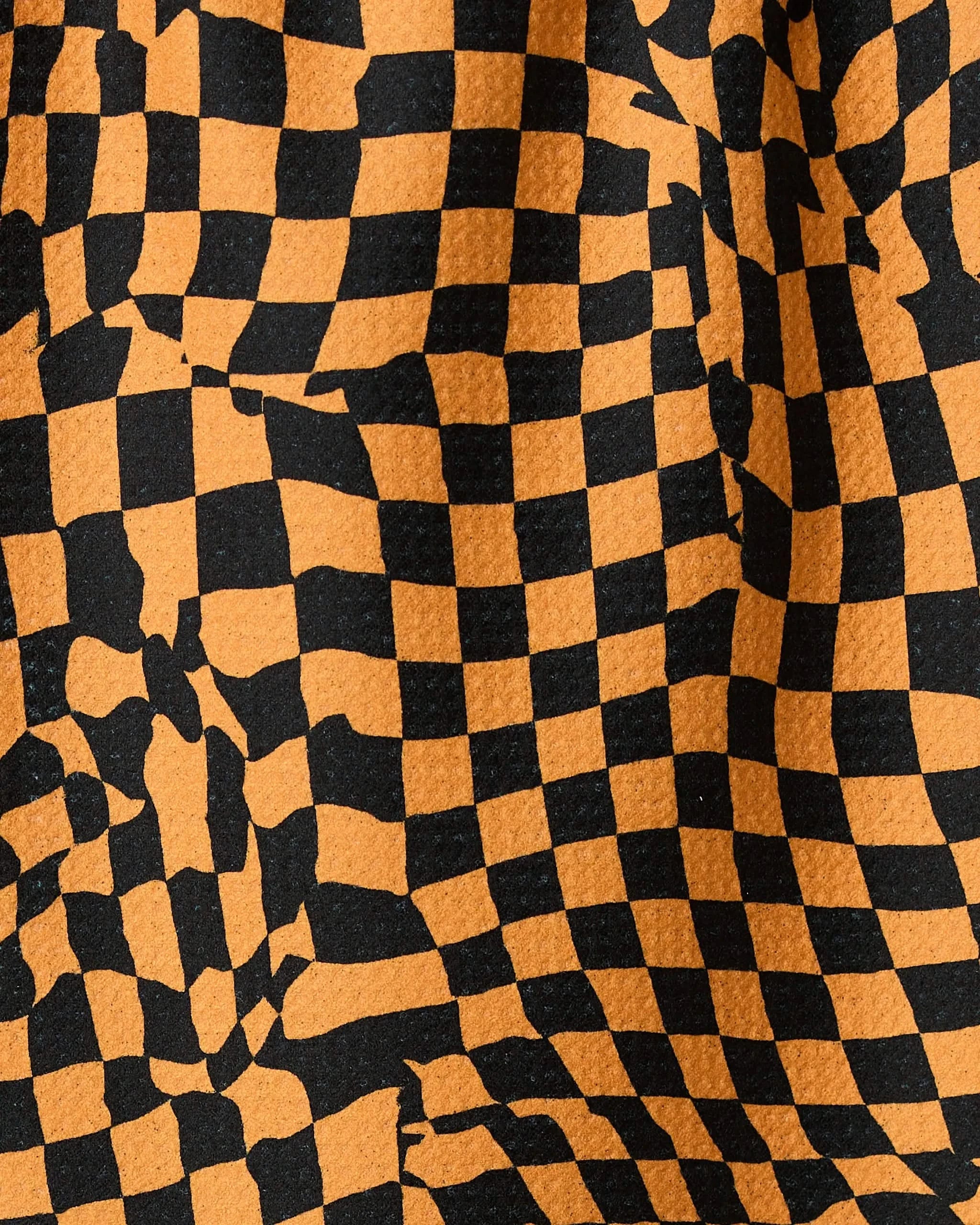 A close-up of the Geometry Glitchy Check Orange recycled kitchen towel shows its orange and black checkered design with a wavy, optical illusion pattern and a textured, rippled surface.