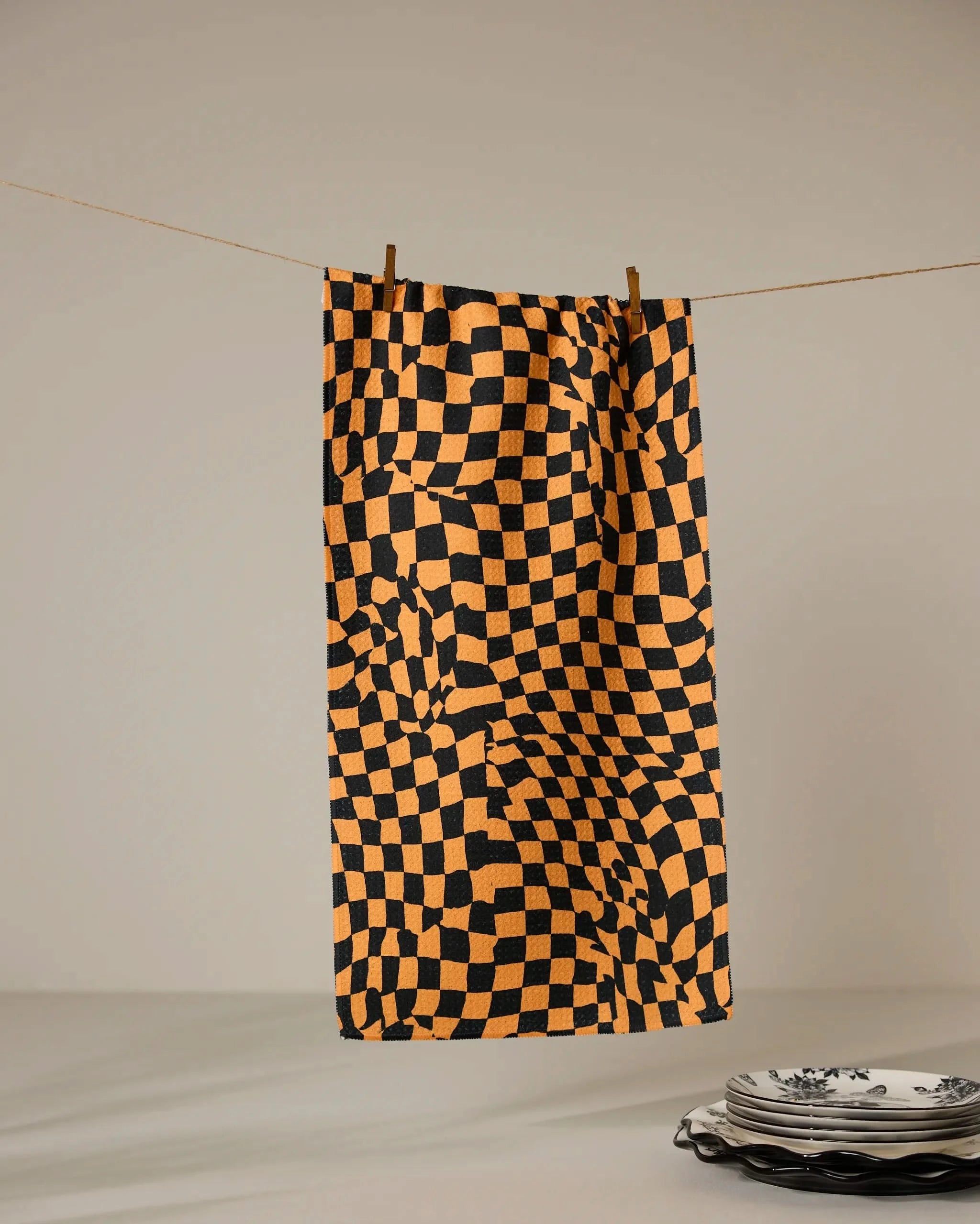 The Geometry Glitchy Check Orange ultra-absorbent towel, featuring a warped orange and black checkerboard pattern, hangs from a string by two clothespins, with white plates adorned in blue designs stacked nearby.