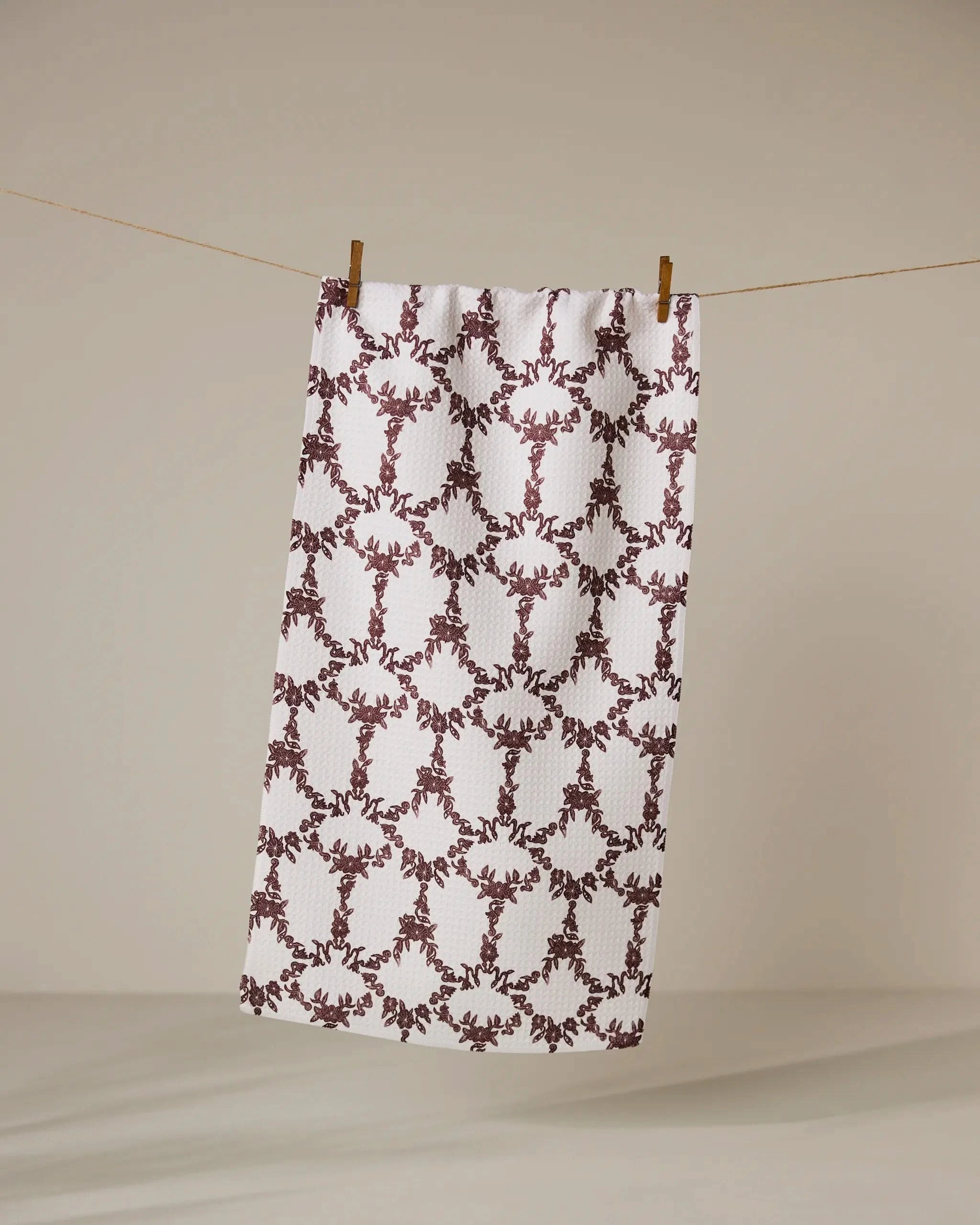 The Geometry Eye on You Floral bar towel, featuring a white base with maroon floral and geometric designs, hangs from a clothesline with two wooden clothespins against a plain beige background.