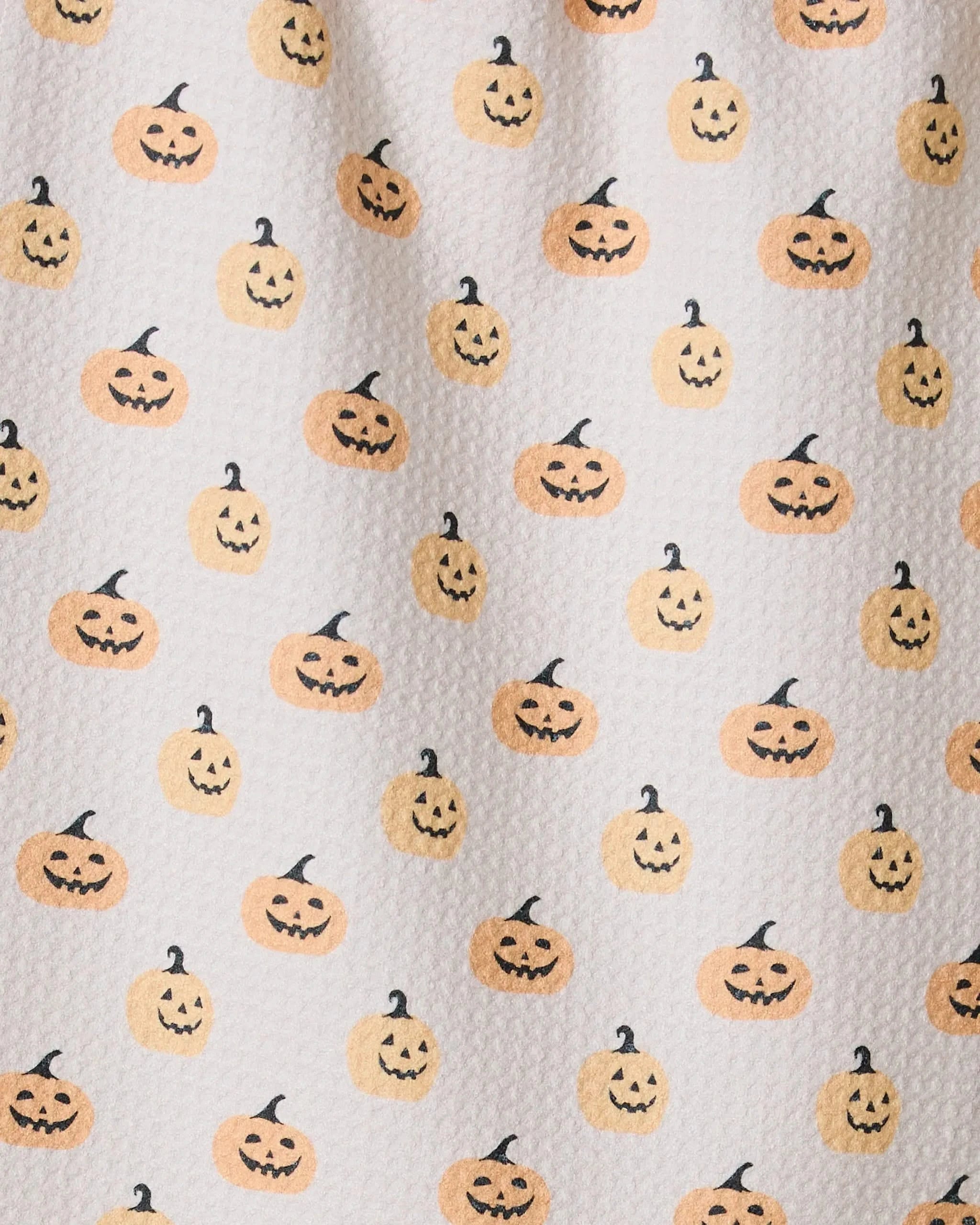 The Geometry Spice and Smiles bar towel is white, textured, and made from recycled materials, featuring a repeating pattern of smiling orange jack-o-lanterns with black stems.