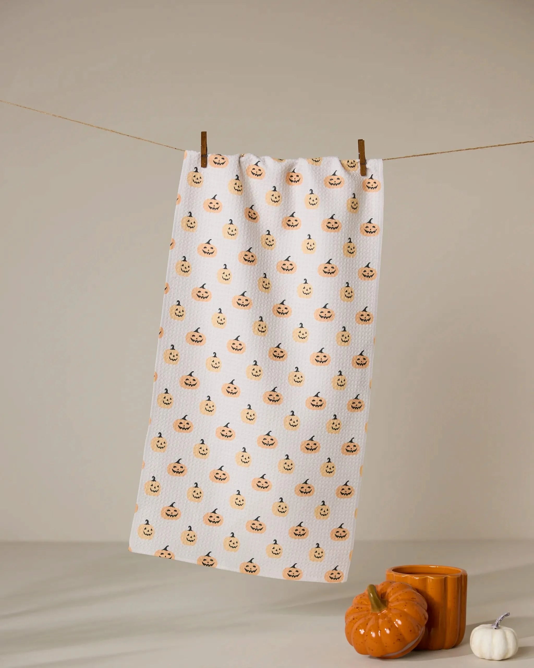 The Geometry Spice and Smiles dish towel, featuring a white ultra-absorbent fabric with smiling pumpkin faces, hangs from a clothesline above an orange mug, ceramic pumpkin, and small white pumpkin.