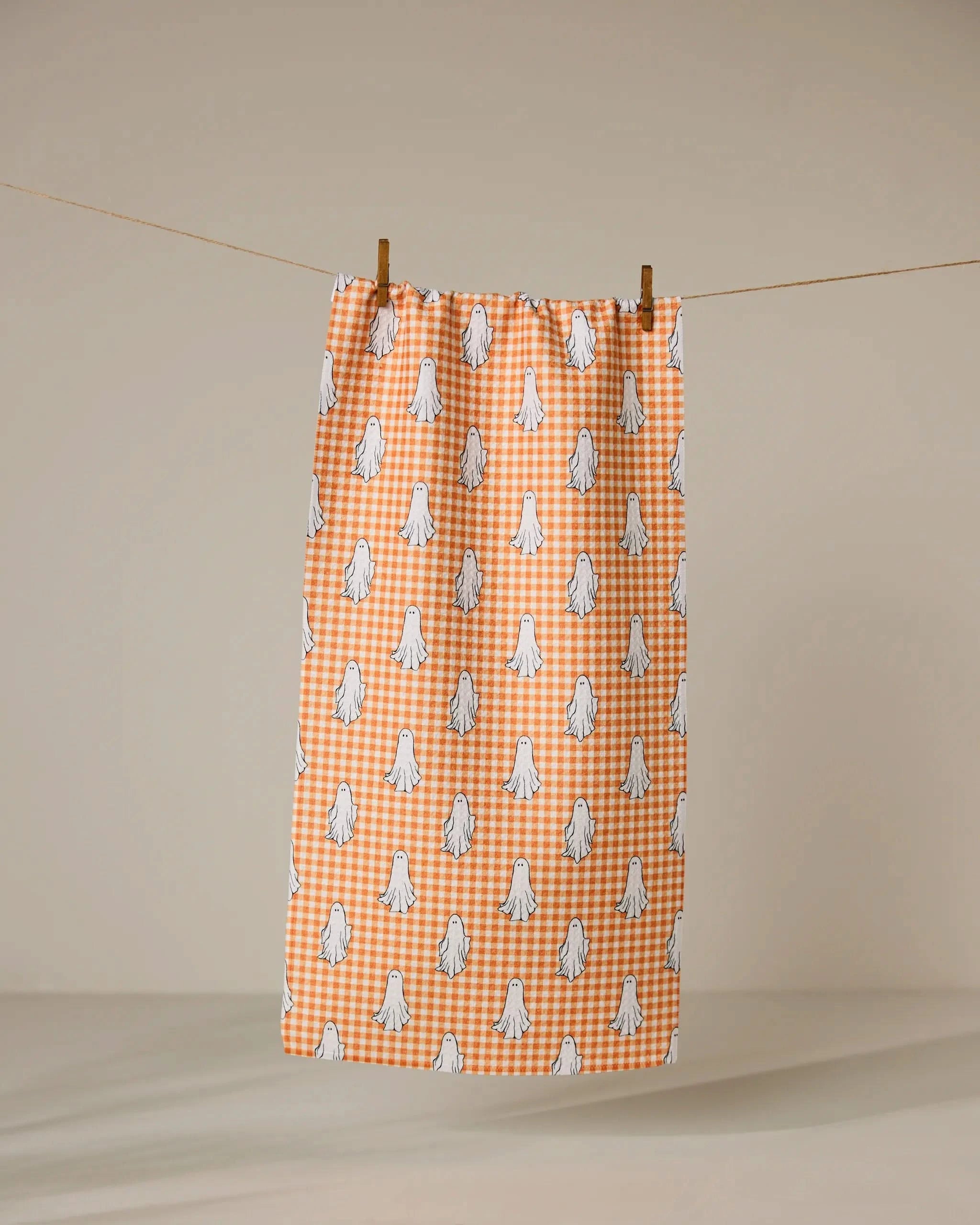 The Gingham Ghosts bar towel by Geometry, featuring orange and white checks with small ghost patterns, hangs on a clothesline with two wooden clothespins against a plain beige background.