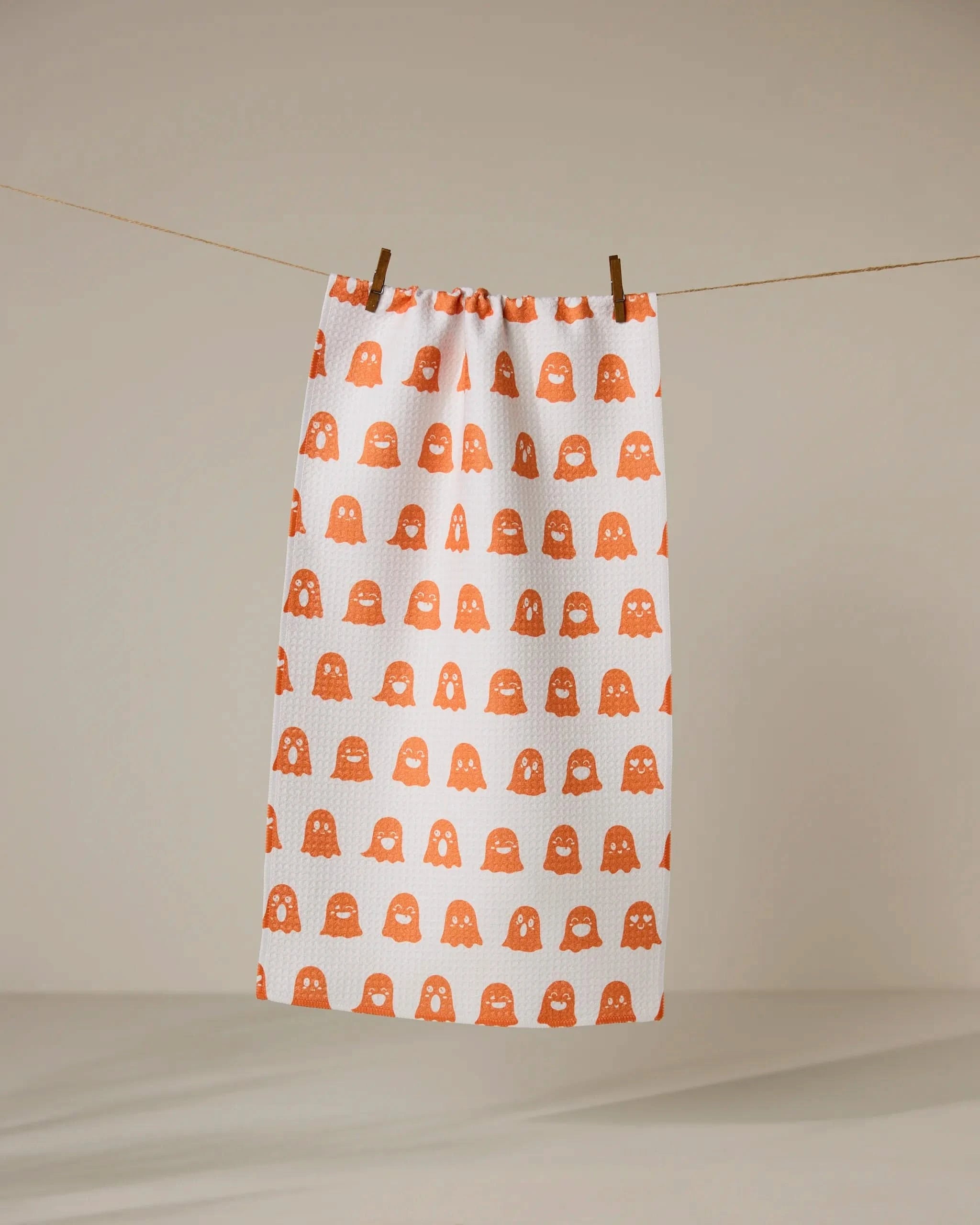 The Geometry Emotional Boos towel, white with an orange pattern of smiling ghosts, hangs from a clothesline with two wooden clothespins against a neutral background.