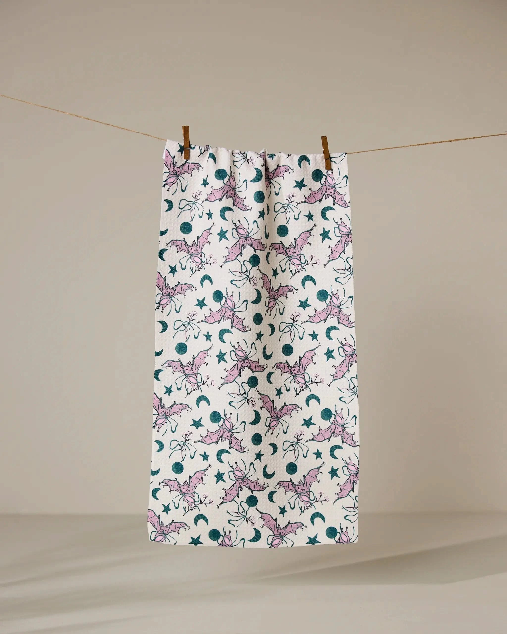 A white recycled kitchen towel by Geometry, called Bats with Bows, hangs on a clothesline against a neutral background. It features a repeating pattern of bats with bows in purple and dark teal crescent moons and stars.