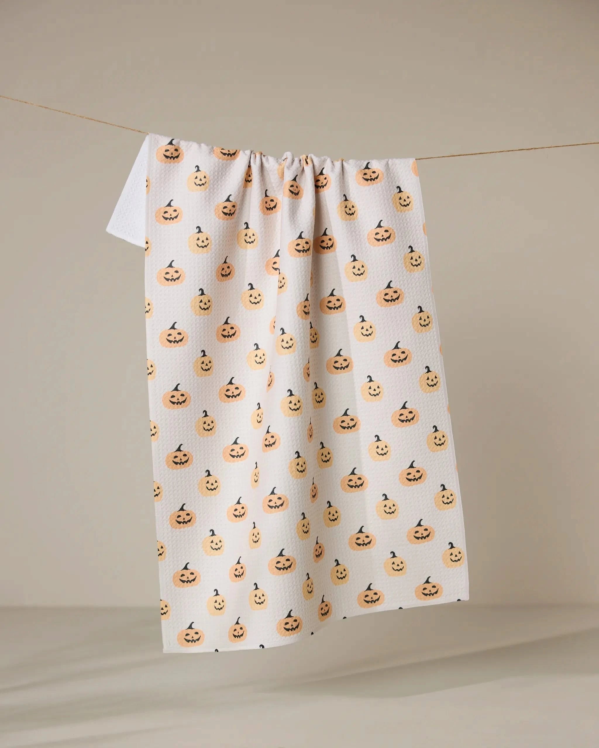 The Spices and Smiles tea towel by Geometry, featuring ultra-absorbent white fabric with cheerful orange jack-o-lanterns, hangs from a clothesline against a beige background.