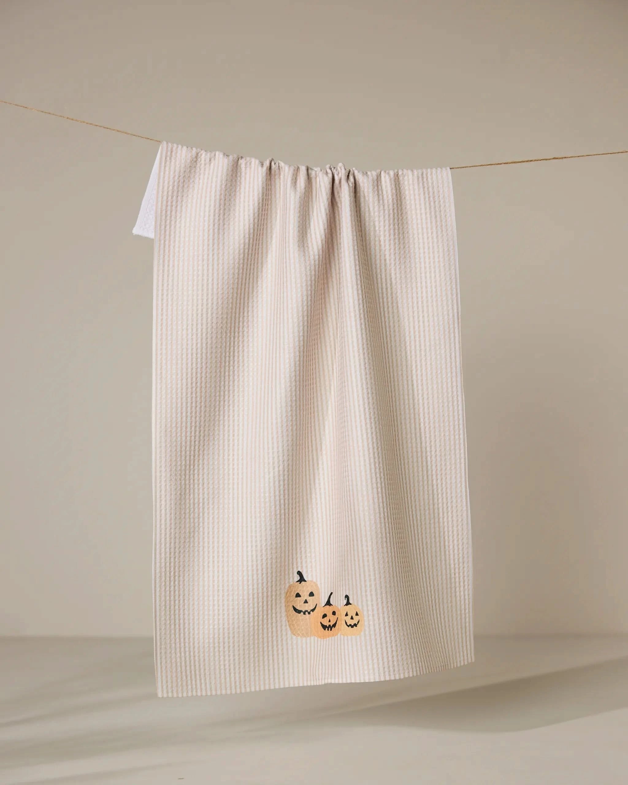 The Geometry Gourdgeous tea towel features beige and white stripes, recycled material, and three small orange jack-o’-lanterns embroidered at the bottom, shown hanging against a plain background.