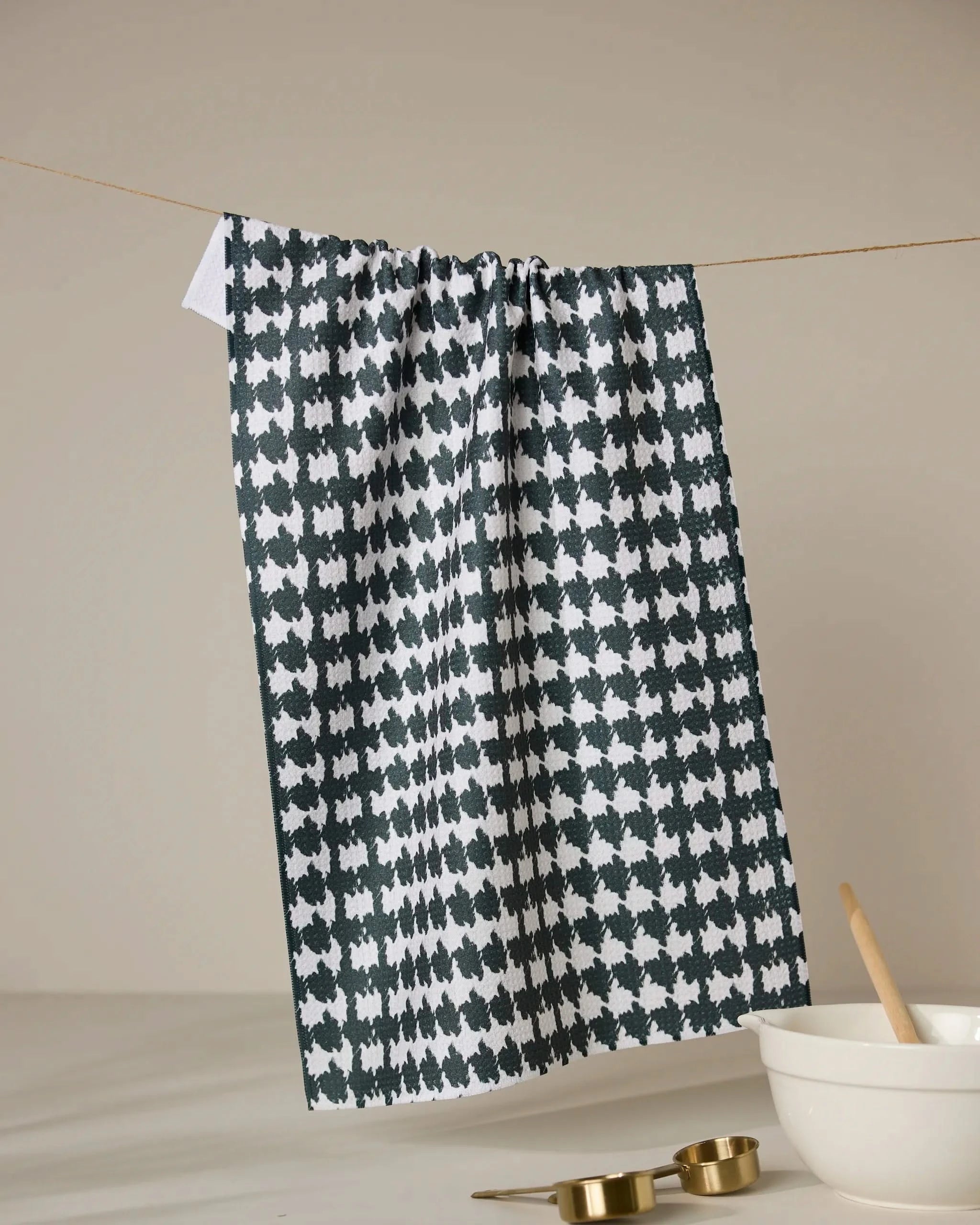 The Geometry Electric Houndstooth Black tea towel, made from recycled materials, hangs on a clothesline above a white surface with a white mixing bowl, wooden spoon, and gold measuring cups nearby.