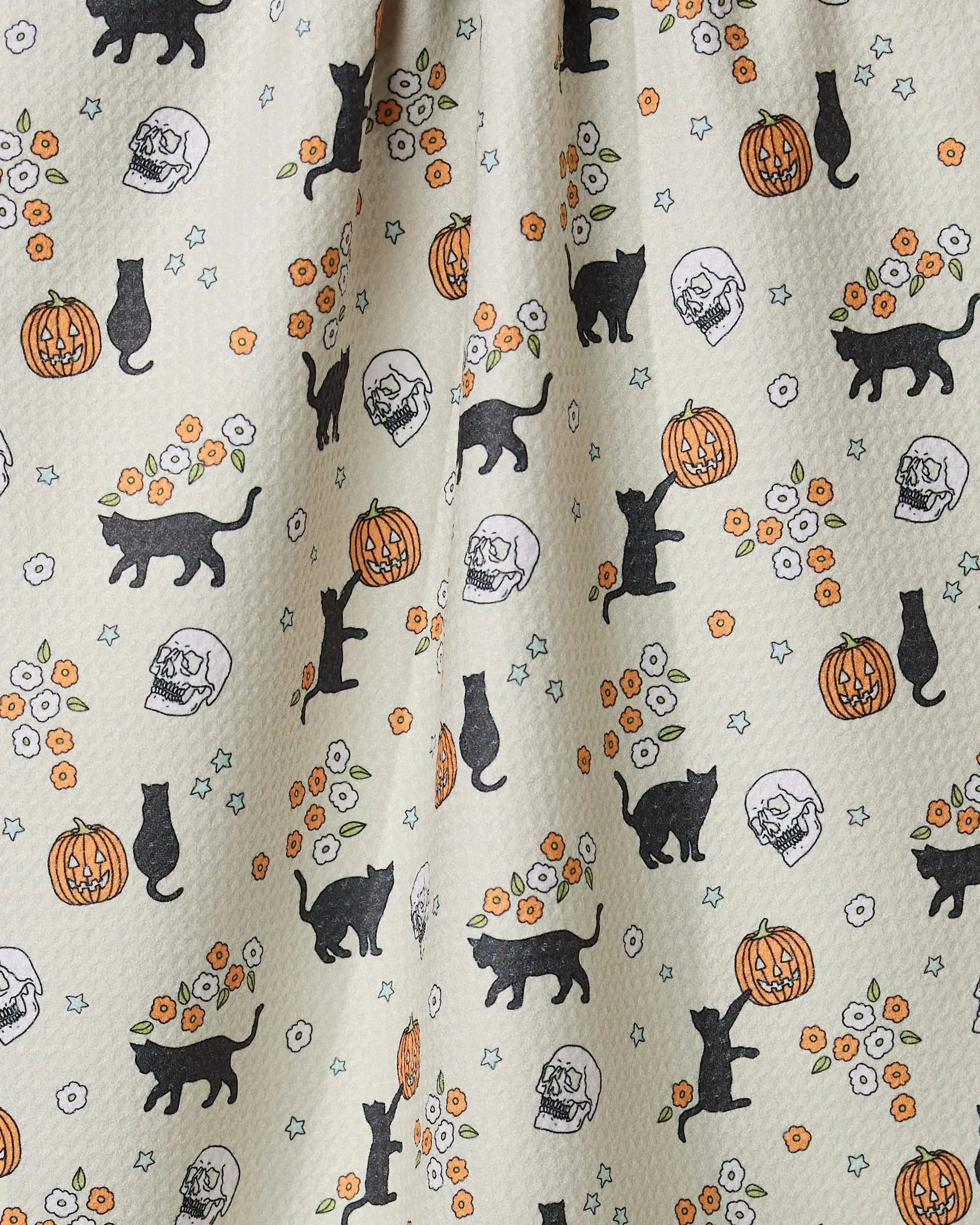 The Geometry Spooky Parade tea towel features black cats, pumpkins, skulls with flowers, and star accents on a light background—evoking Halloween vibes. Made from recycled materials.