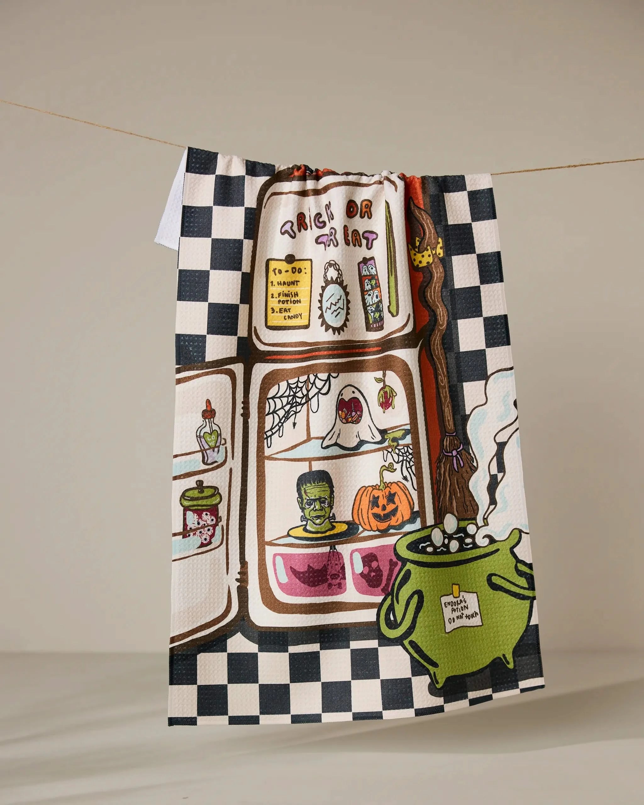 Geometry’s “Spooky Roommates” eco-friendly kitchen towel features a checkered border, spooky potions, a green bubbling cauldron, Frankenstein head, pumpkins, cobwebs, and “Trick or Treat” at the top.
