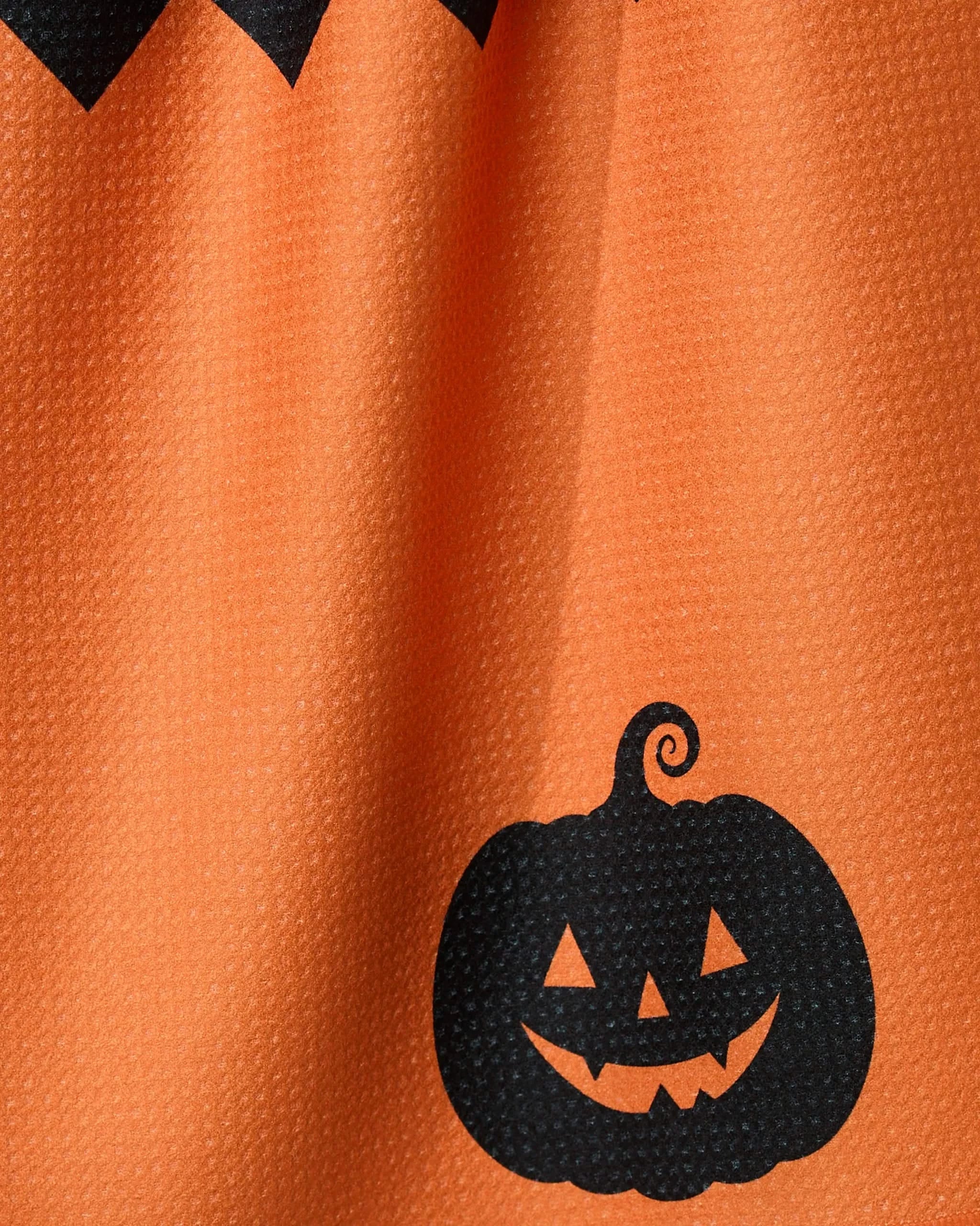 Close-up of Geometrys Spooky Split recycled kitchen towel with an orange texture, black zigzag top border, and a smiling Jack-o’-lantern near the bottom right—perfect for adding Halloween cheer to your kitchen.