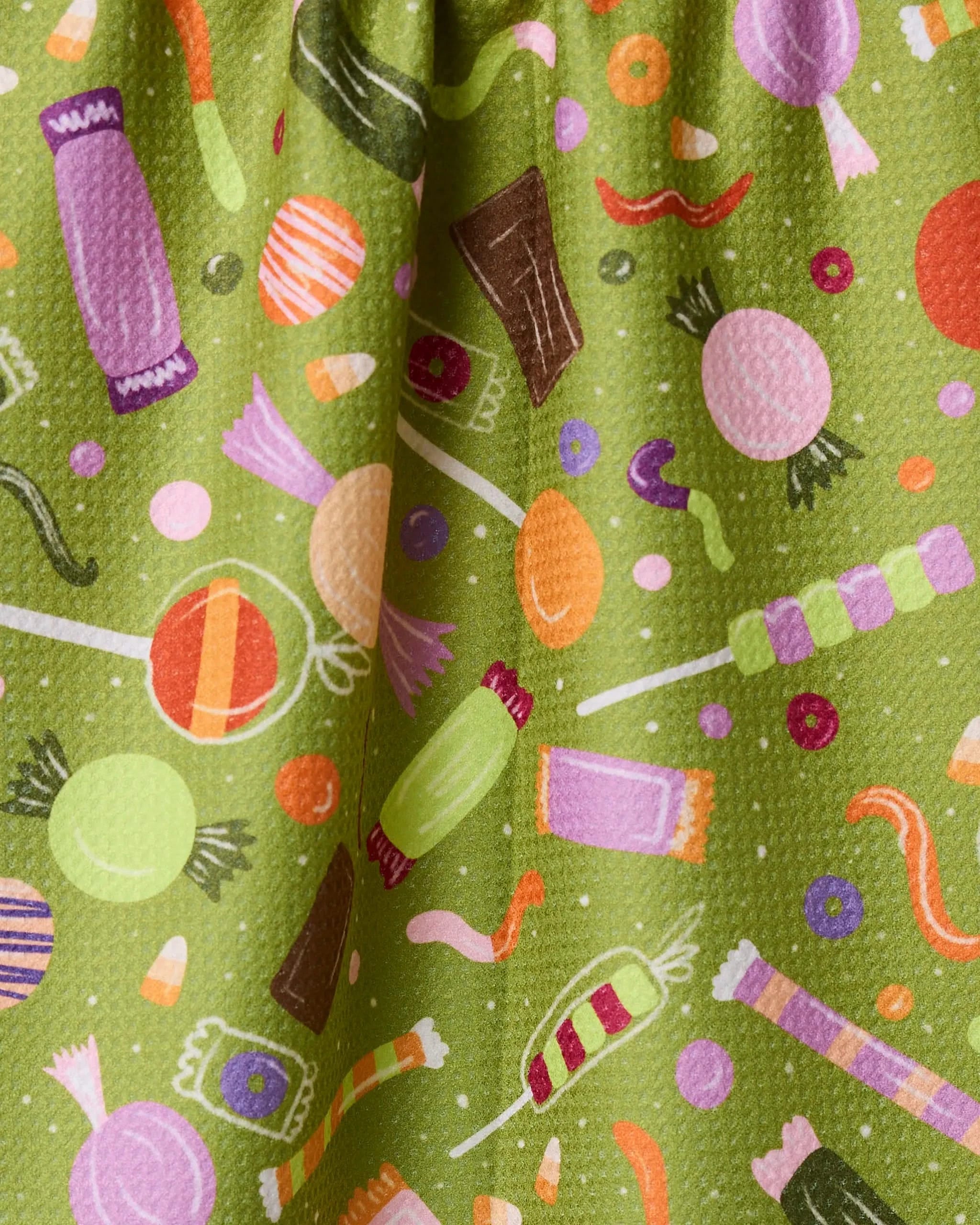 Geometry’s Sweet Treasures tea towel is crafted from recycled materials and features a vibrant green fabric adorned with lollipops, wrapped candies, chocolate bars, candy corn, and gummy worms.