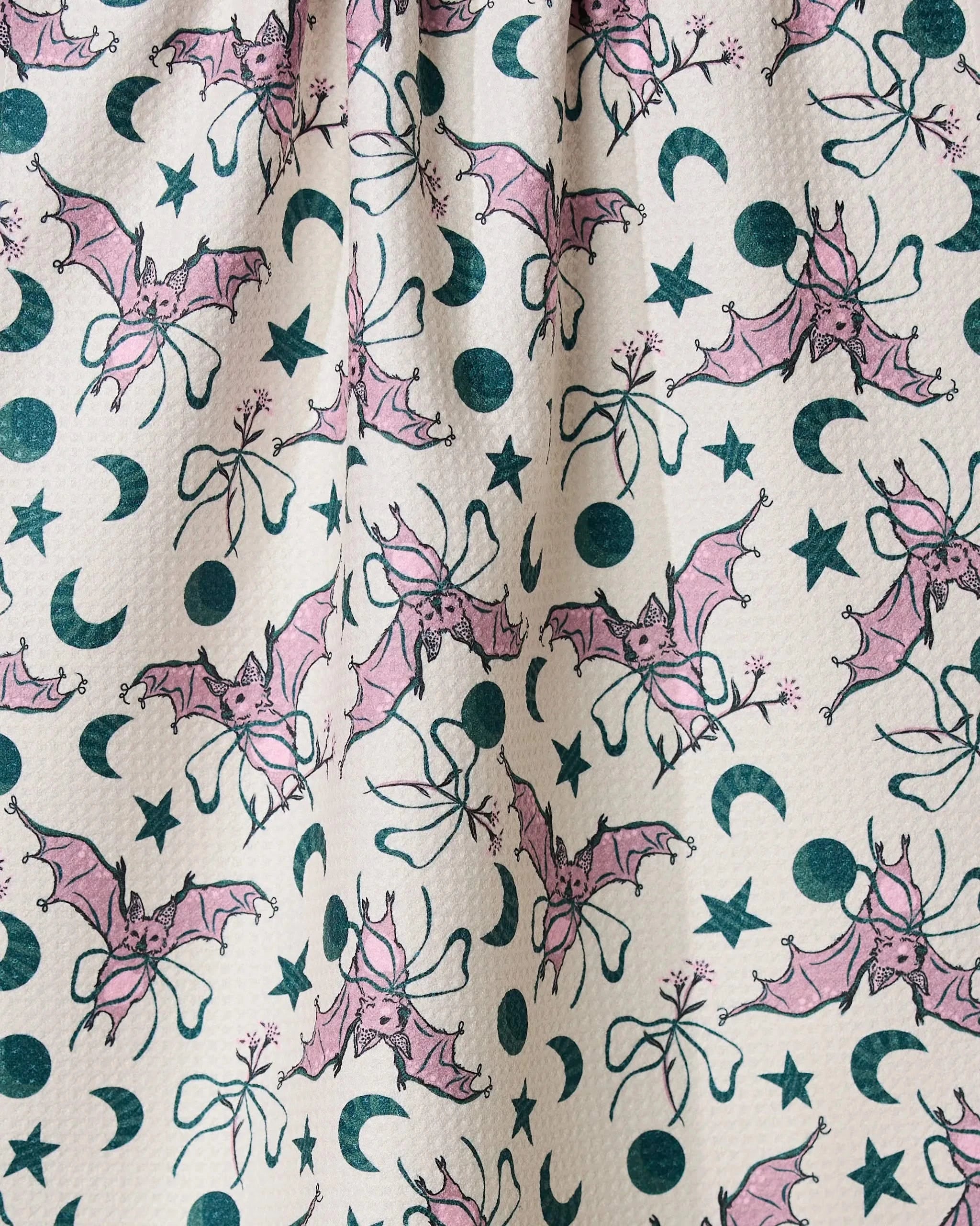 The Geometry Bats With Bows tea towel features pink bats, green moons, stars, and bows on white. Made from recycled materials, its whimsical design is quick-drying with a slightly wrinkled texture for daily use.