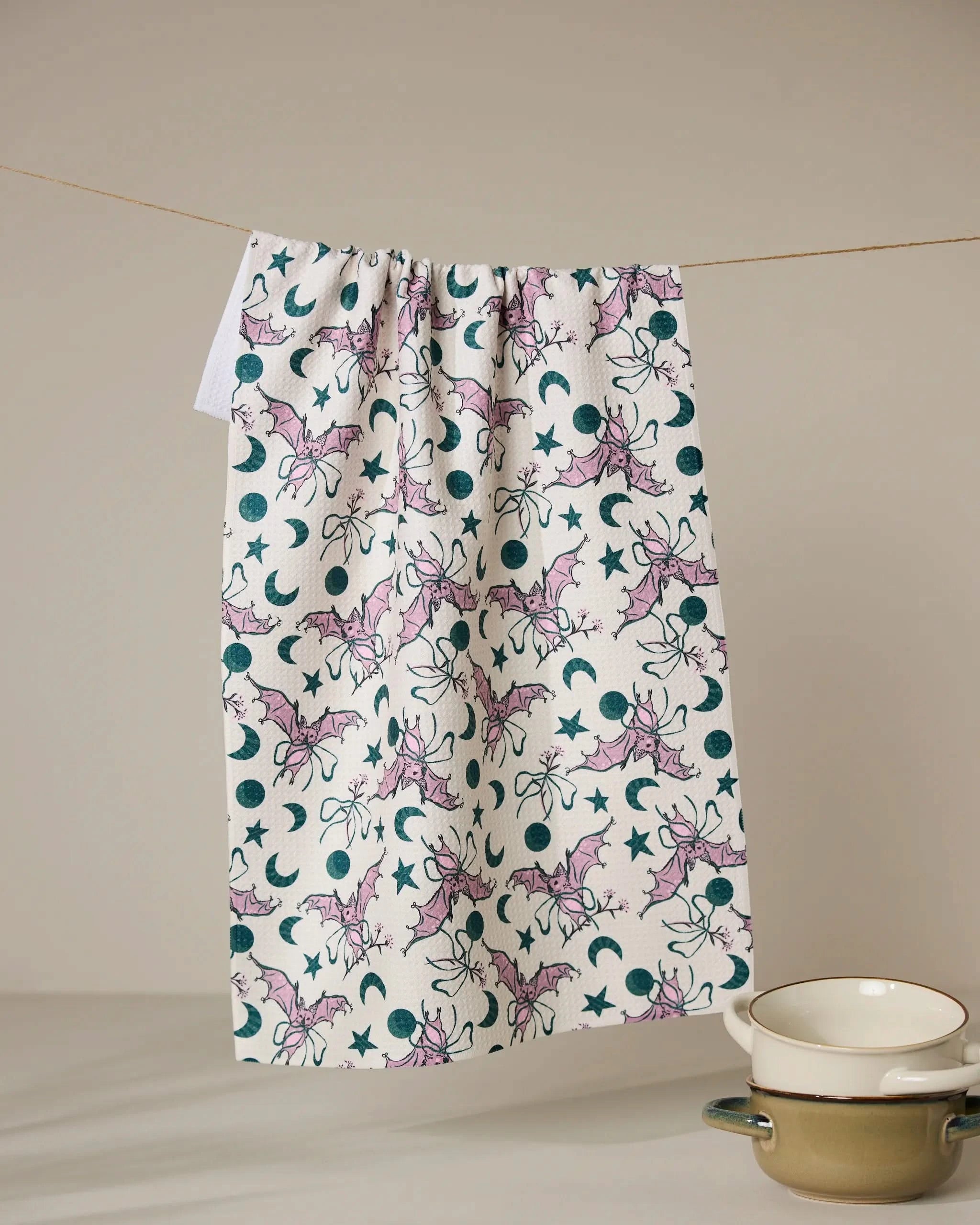 The Geometry Bats With Bows quick-drying tea towel, featuring a whimsical pattern of purple bats, moons, and stars, hangs on a clothesline above two stacked white bowls against a neutral background.