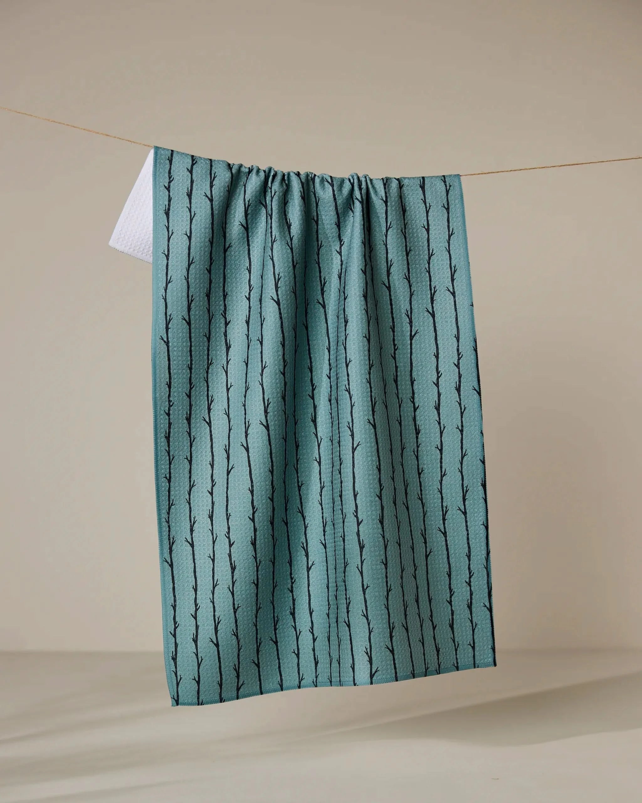 The Geometry Branch Stripes tea towel in teal with a black branch pattern, made from recycled materials, hangs on a clothesline against a neutral beige background.