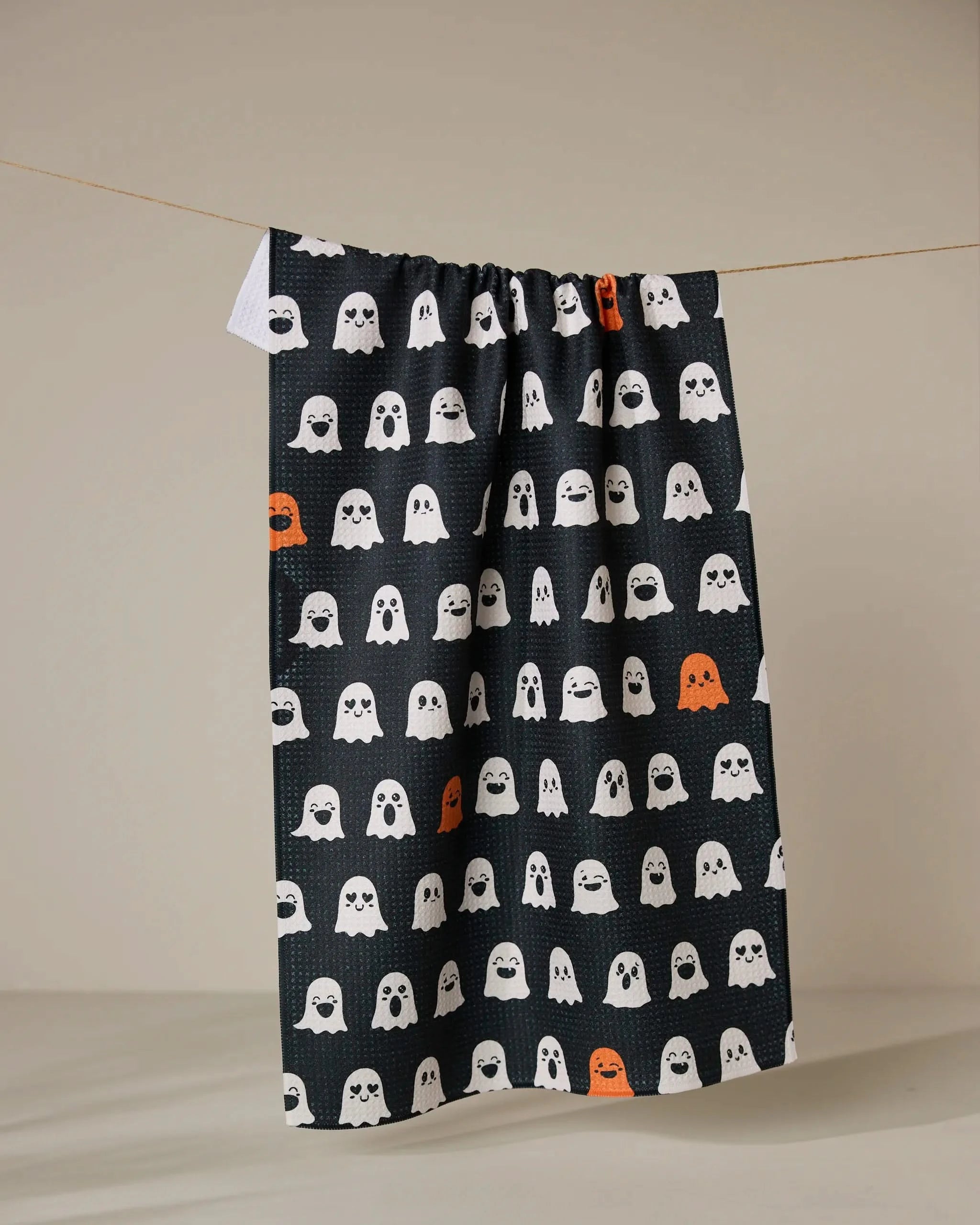 The Geometry Emotional Boos tea towel, featuring quick-drying material and a pattern of mostly white and some orange cartoon ghosts, hangs from a string against a plain beige background.